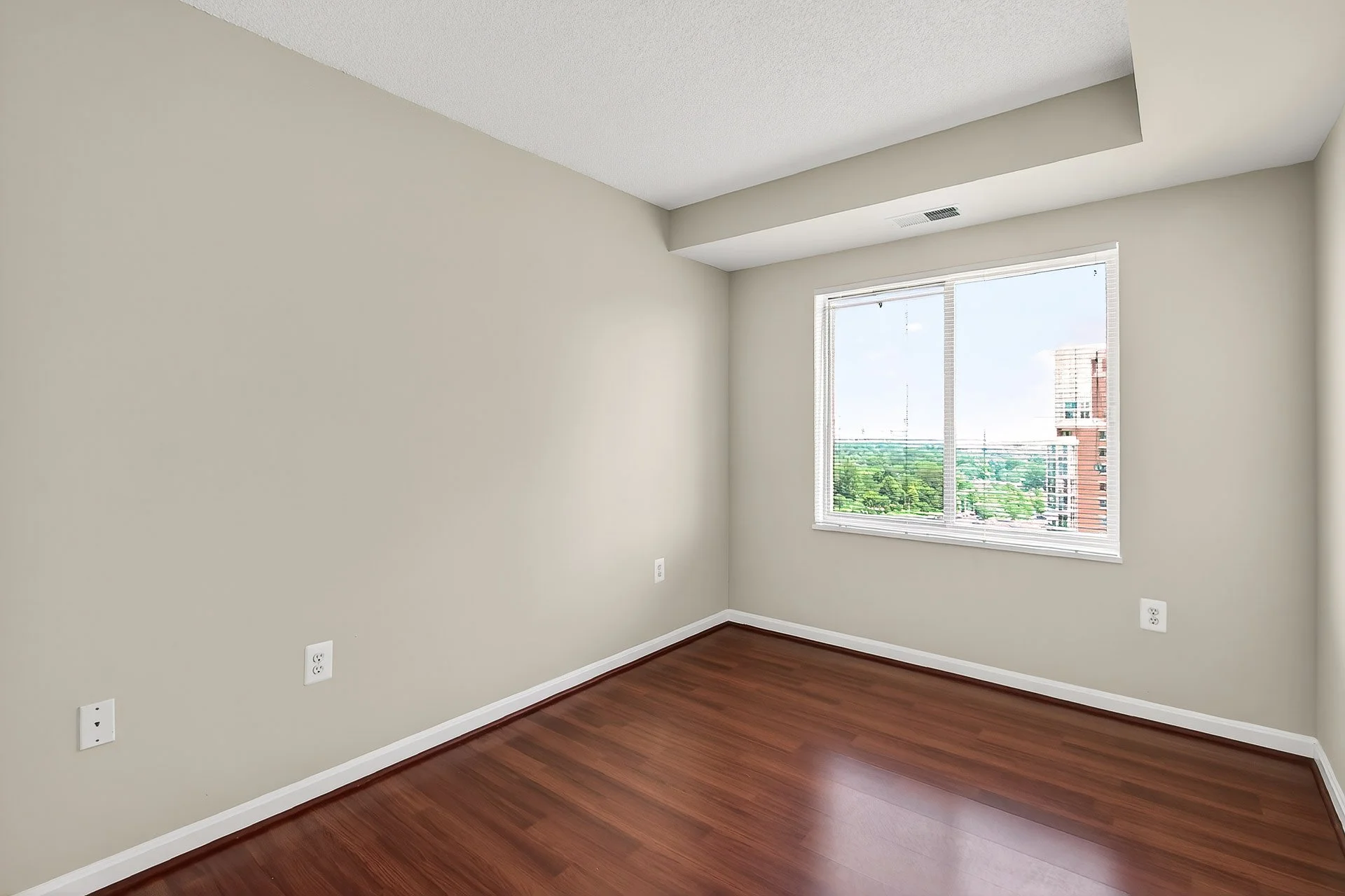 Empty room with hardwood floors, white walls, a large window showing a cityscape and green trees, and electrical outlets on the wall.