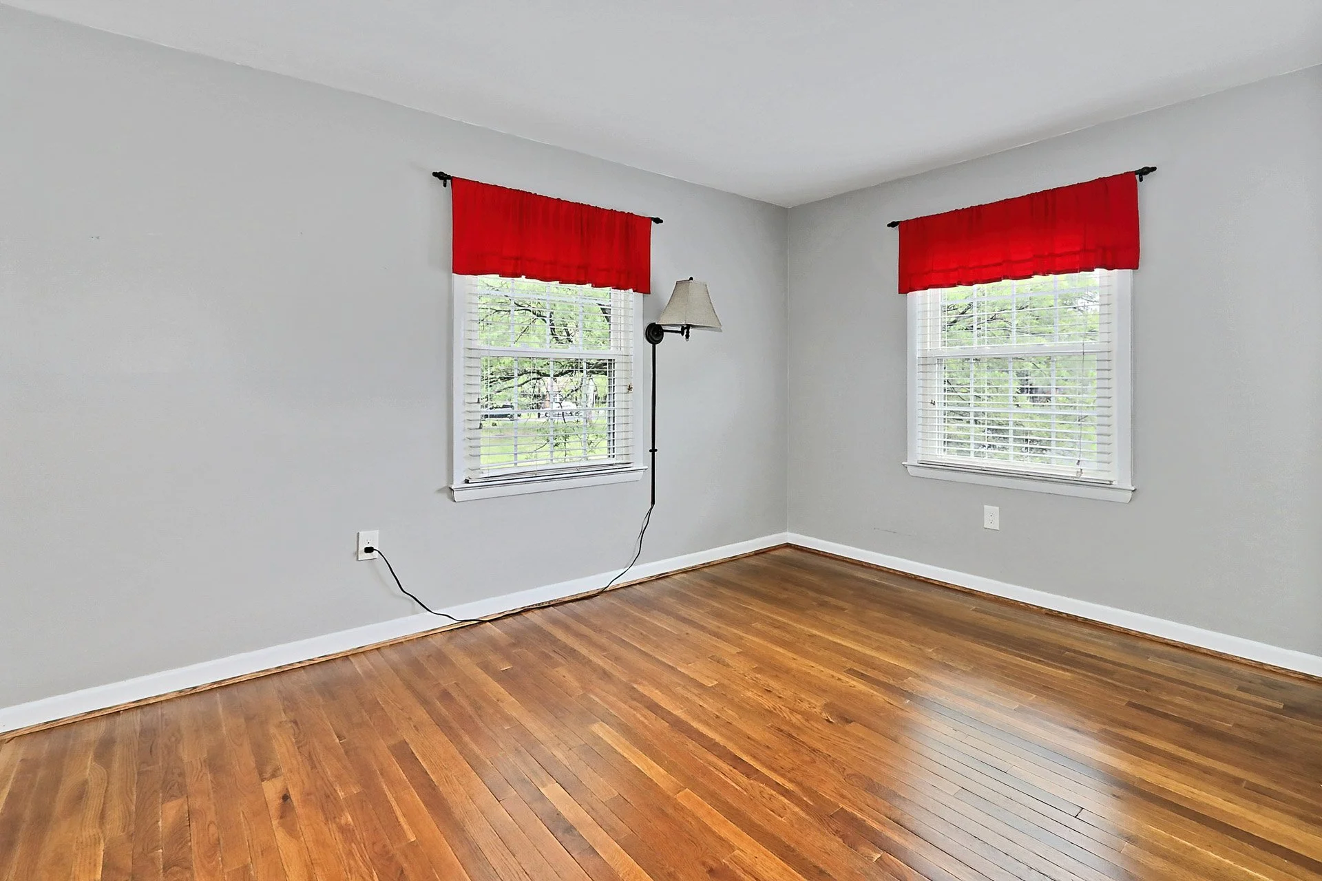 Empty room with hardwood floors, two windows with white blinds and red valances, a wall-mounted lamp, and an electrical outlet.