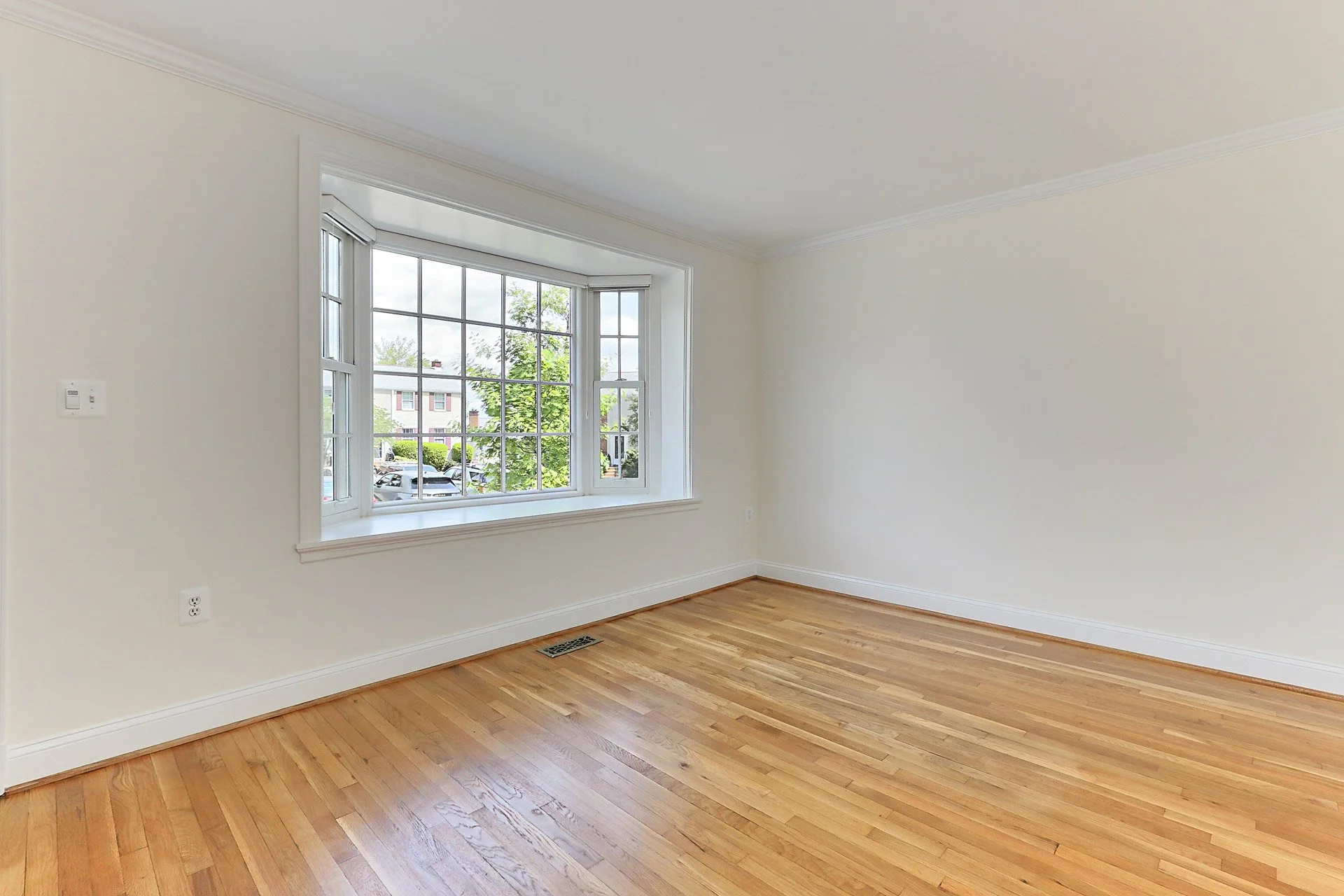 Empty room with hardwood floors, white walls, and a large window showing outside trees and buildings.