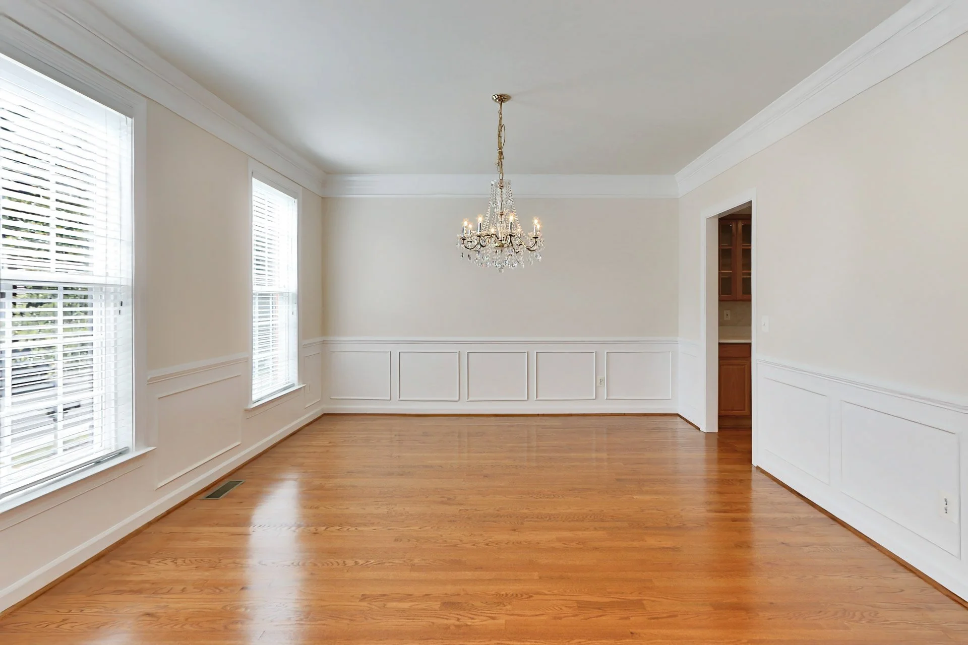 Empty dining room with white walls, hardwood floor, large windows, a chandelier, and a doorway to another room.