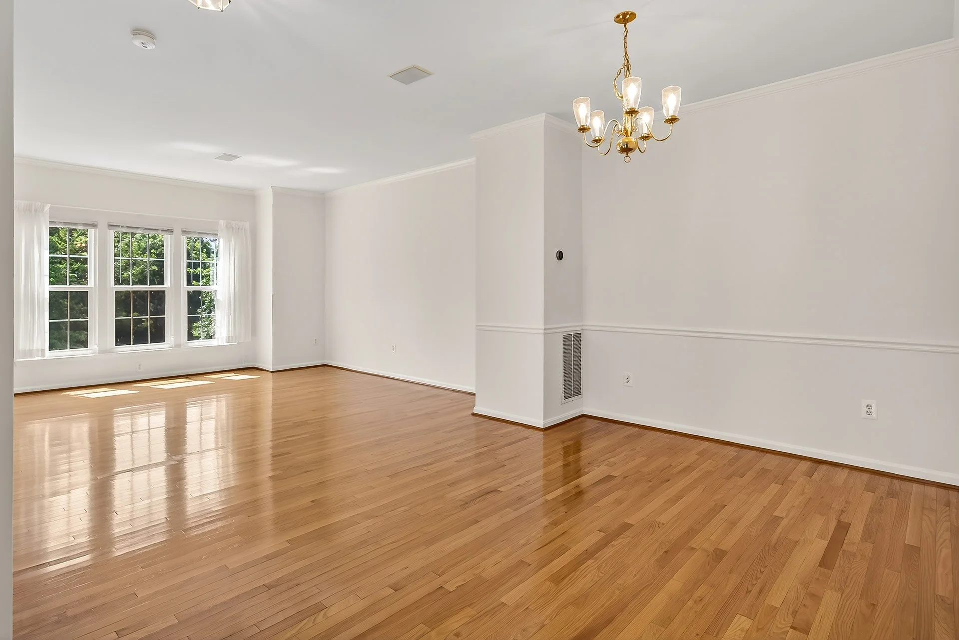 Empty living room with hardwood floors, white walls, large bay window, and a chandelier hanging from the ceiling.