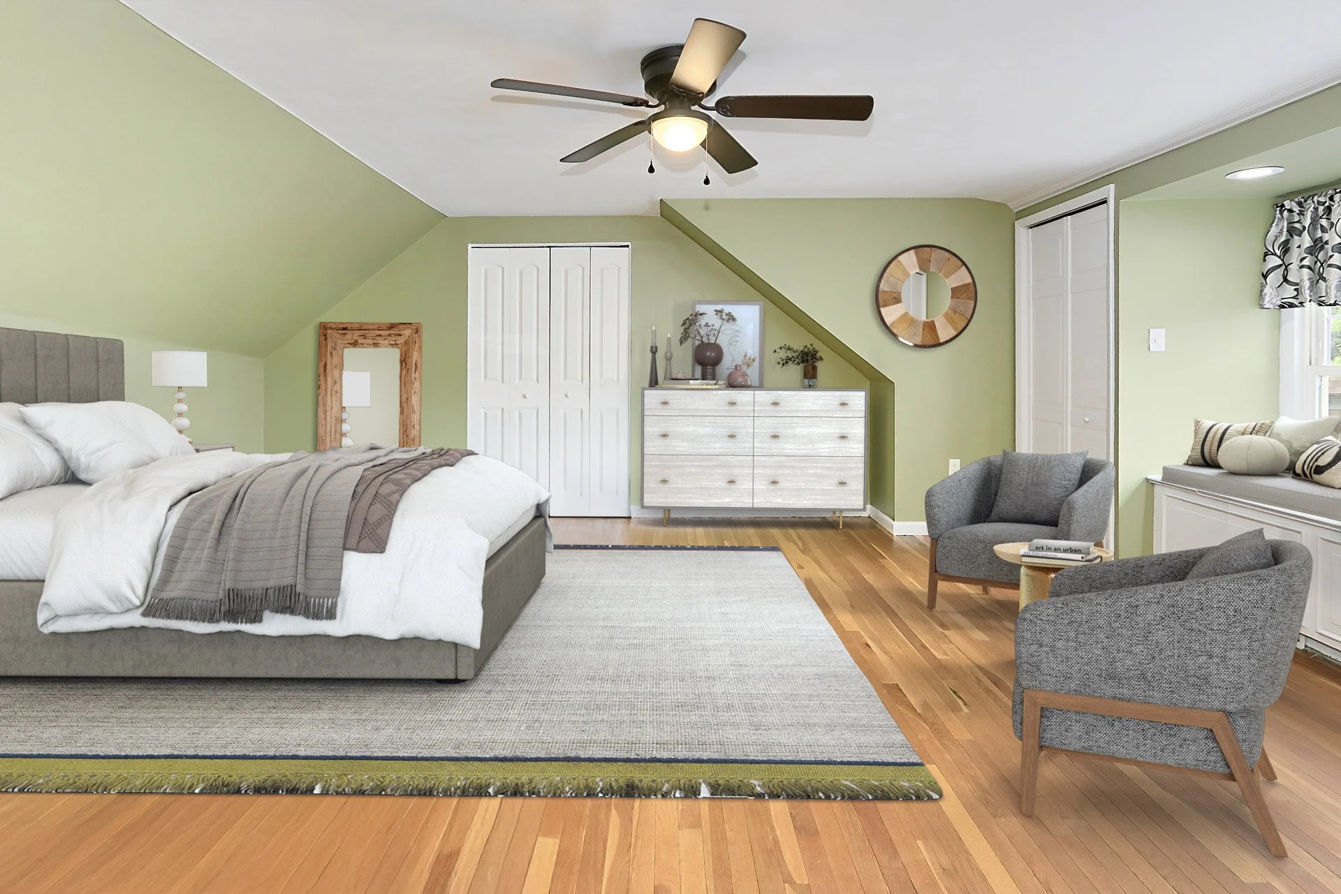A bedroom with a green wall, a ceiling fan, a bed with white bedding and a gray headboard, a small table with a stool, two gray chairs, a window with window seat and cushions, and a white dresser.