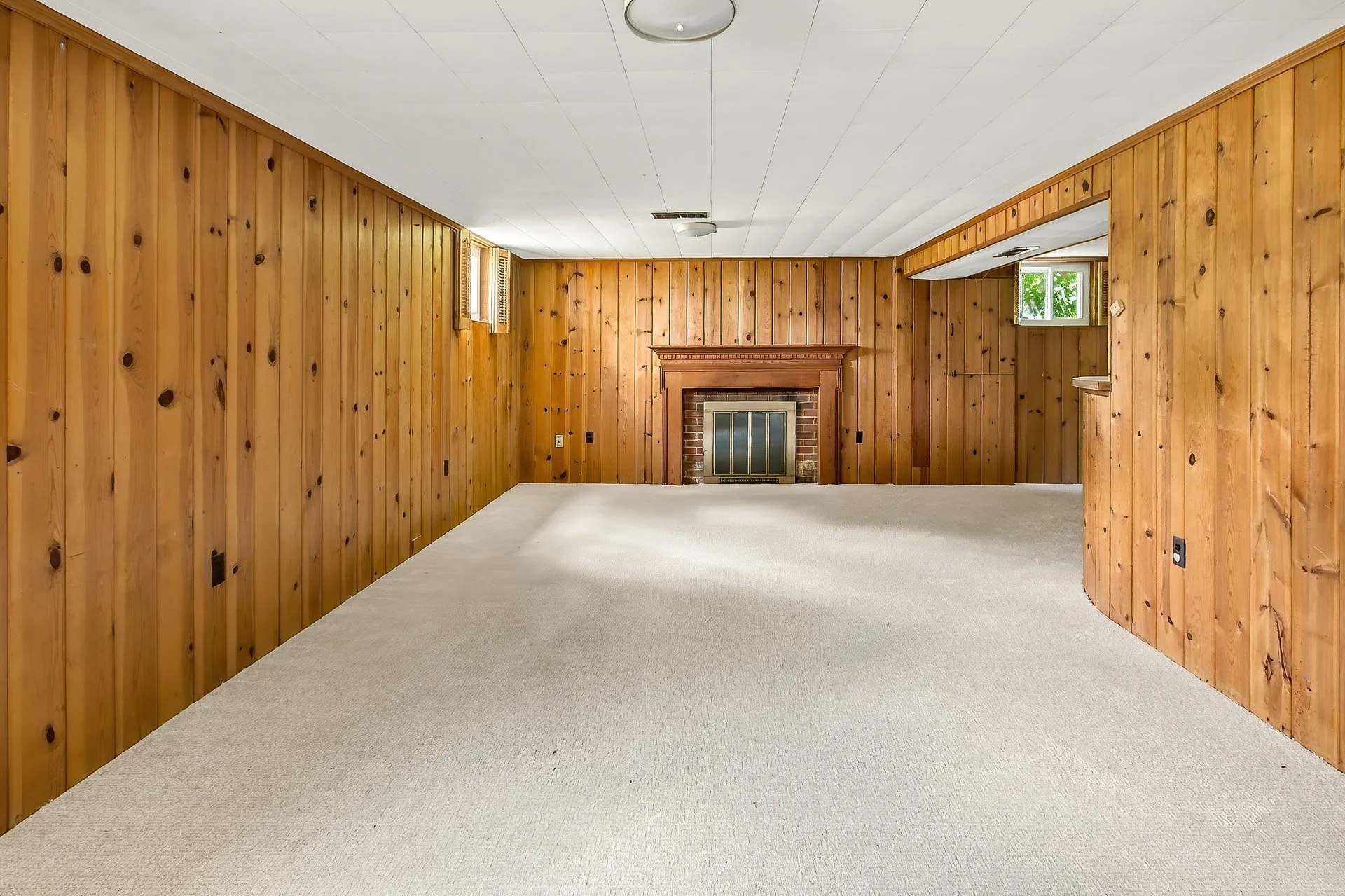 A room with wood-paneled walls, white ceiling, and a fireplace with brick surround and wooden mantel.
