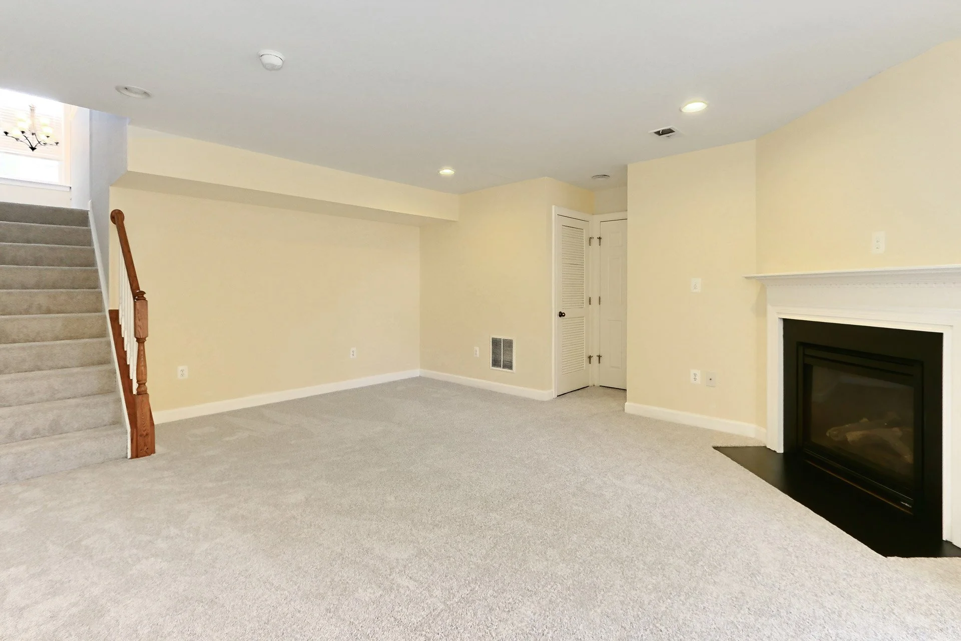 Empty living room with beige walls, a staircase with carpeted steps and wooden handrail, a fireplace with a white mantel, and beige carpet flooring.