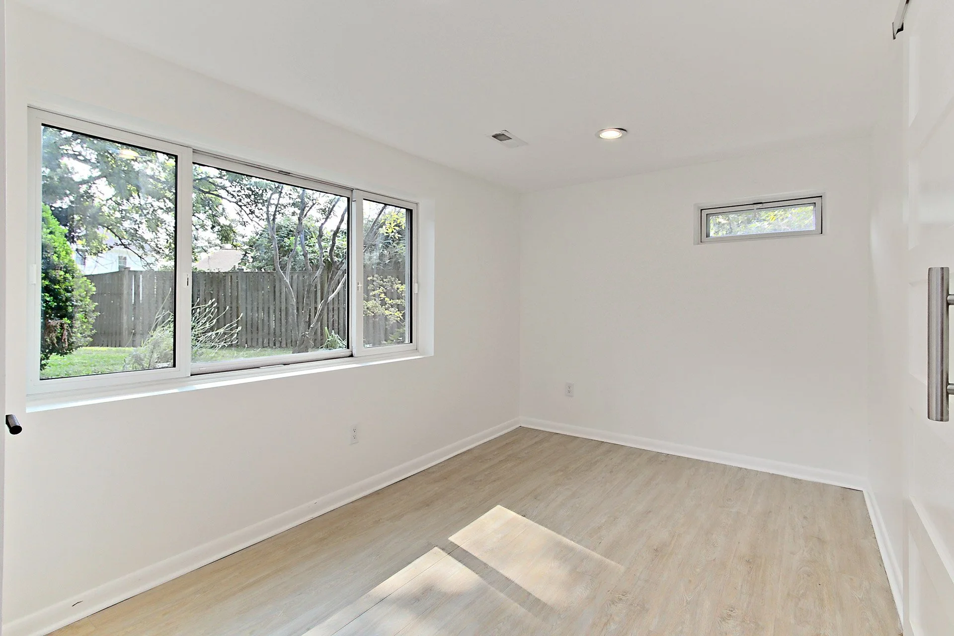 Empty room with large window showing trees and fence outside, white walls, light wood flooring, small horizontal window, and ceiling lights.