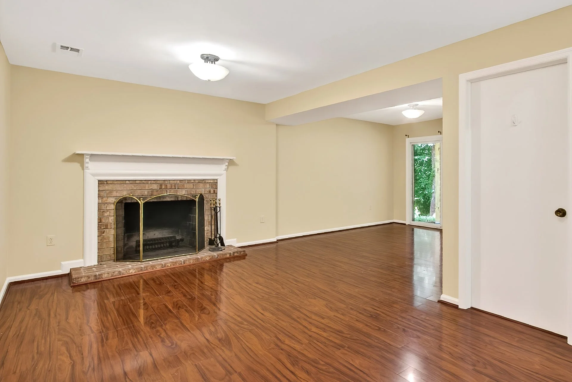 Empty living room with hardwood floors, beige walls, a white brick fireplace with a wooden mantel, and sliding glass door leading outside.