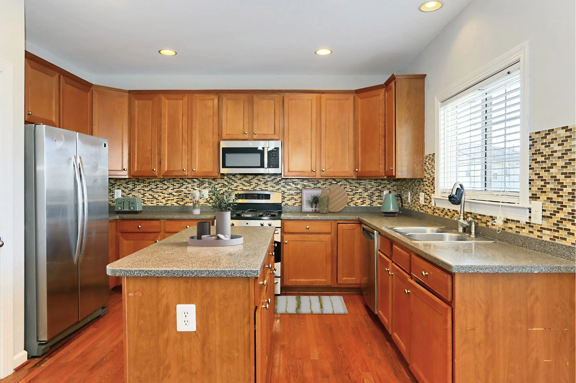 A kitchen with wooden cabinets, a stainless steel refrigerator, a microwave oven, a stove, and a sink beneath a window. There is a small island with a tray and candles, a multicolored mosaic backsplash, and hardwood flooring.