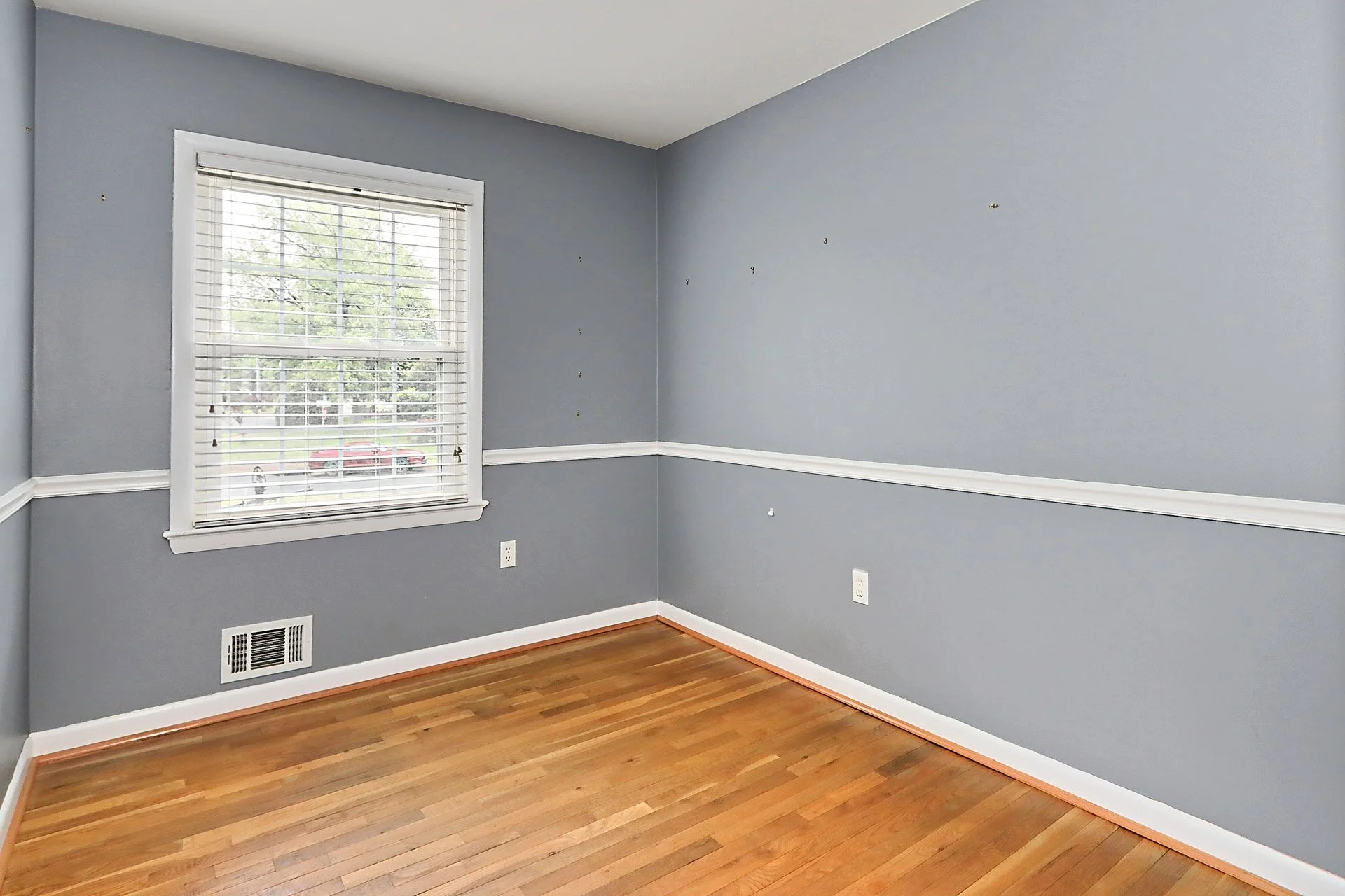 Empty room with gray walls, white trim, wooden floor, and window with blinds.