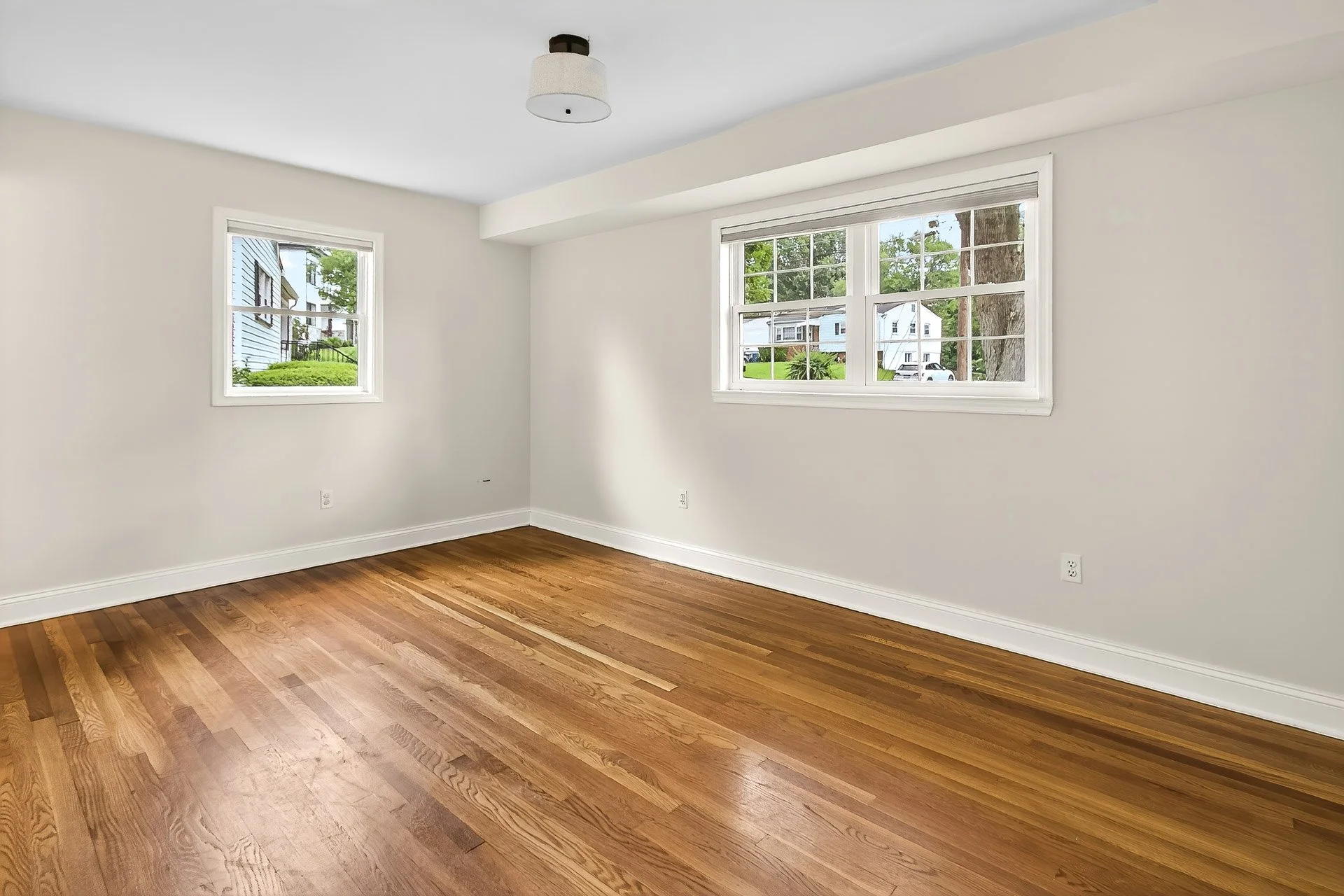 Empty room with hardwood floors, white walls, and two windows showing a residential neighborhood outside.