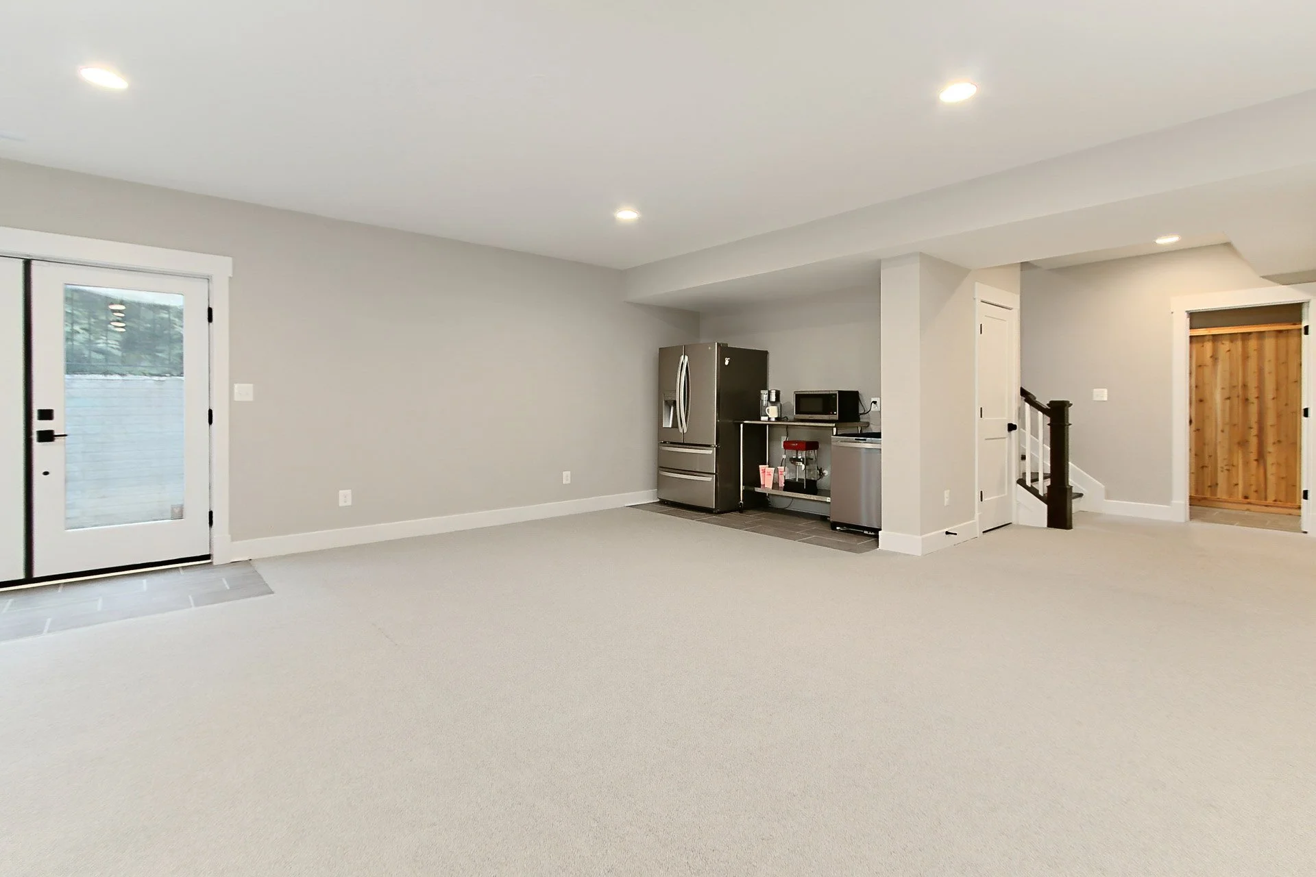 Bright empty basement room with gray walls, beige carpet, and small kitchenette with stainless steel appliances.