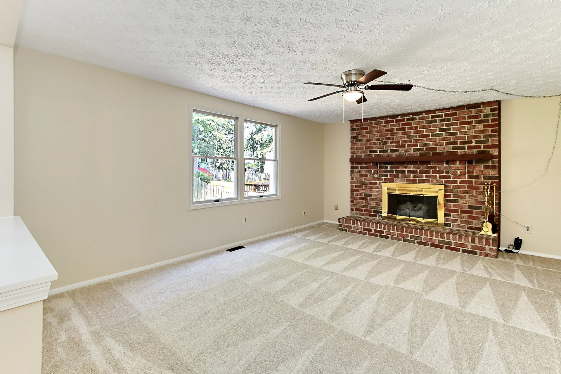 Living room with beige carpet, large window, ceiling fan, and a brick fireplace with a wooden mantle.