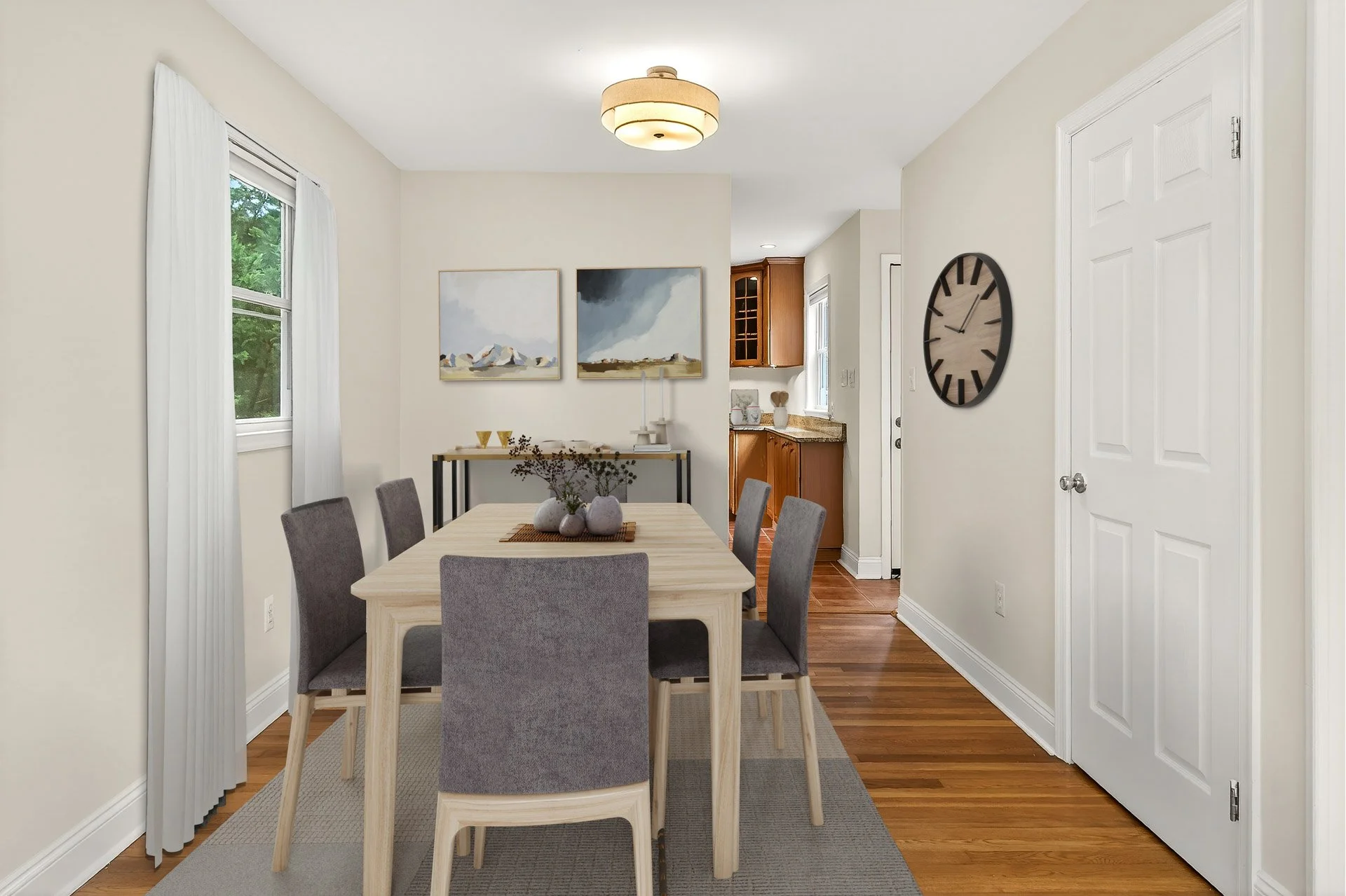 A dining room with a light wood table, six gray upholstered chairs, a gray rug, and minimal decor, with a view of the kitchen in the background and a large wall clock on the right wall.