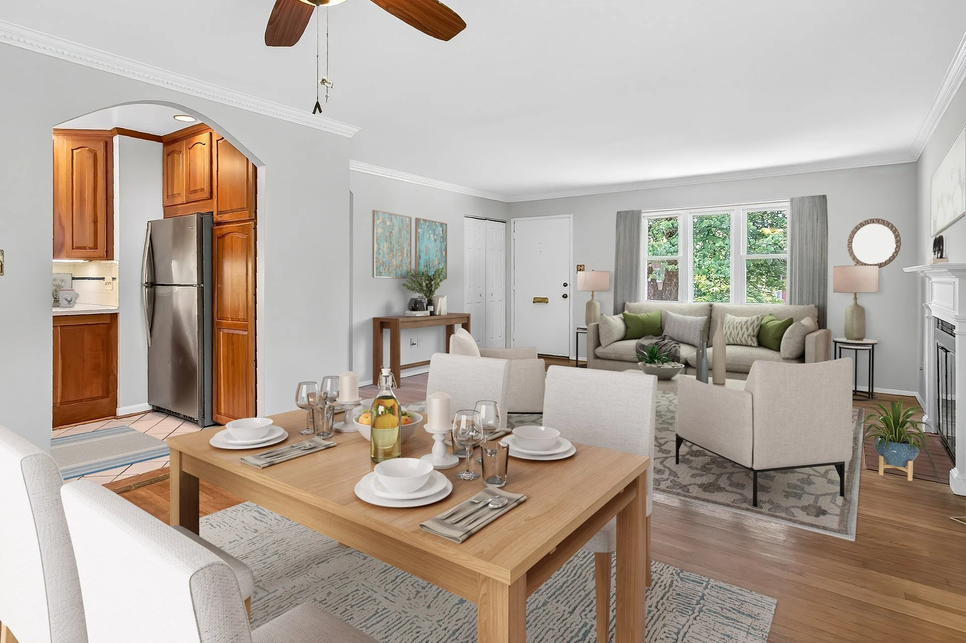 Living and dining room with white walls, hardwood floors, a wooden dining table set for four, and a cozy seating area with sofas and armchairs. The space has large windows with light curtains, a fireplace, a ceiling fan, and decorative night lamps.