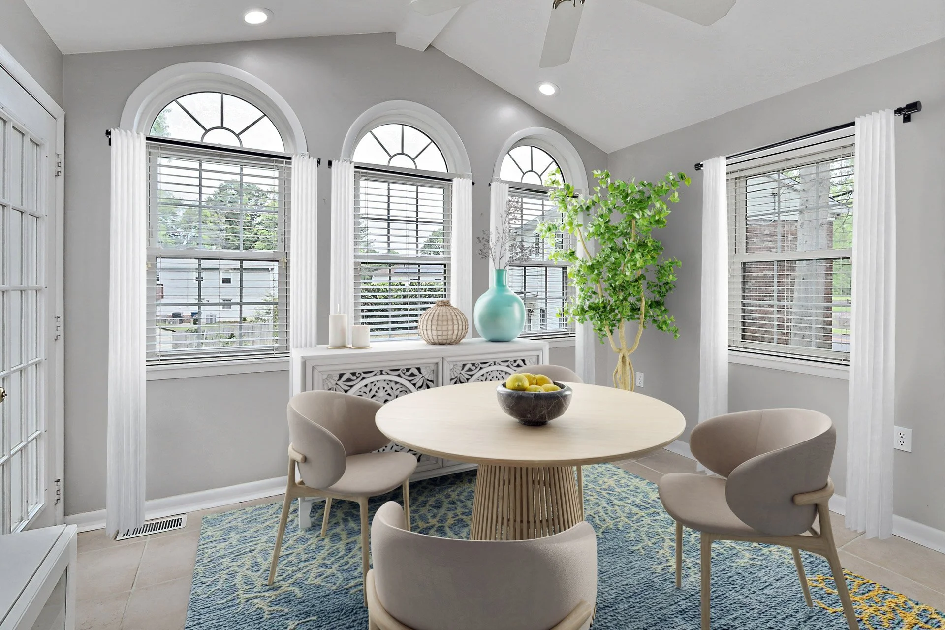 A bright dining room with four beige chairs around a round wooden table. The room has gray walls, large arched windows with white blinds, and a blue patterned rug. Decor includes a large turquoise vase with branches, a woven decorative object, candle