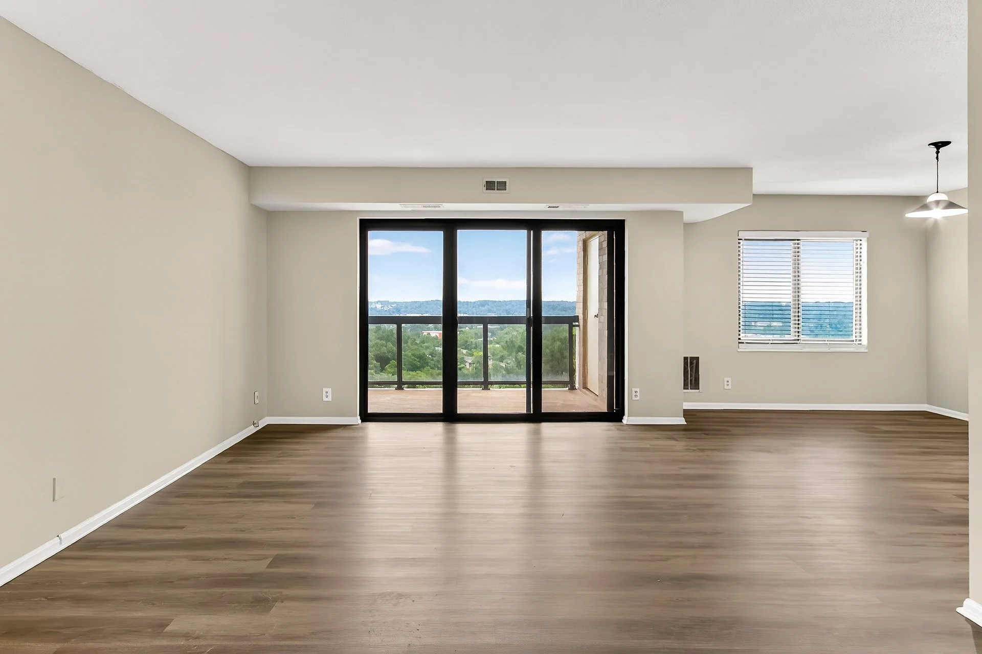 Empty living room with wooden flooring, large sliding glass door leading to a balcony, and window with blinds; neutral-colored walls and ceiling, with a hanging light fixture.