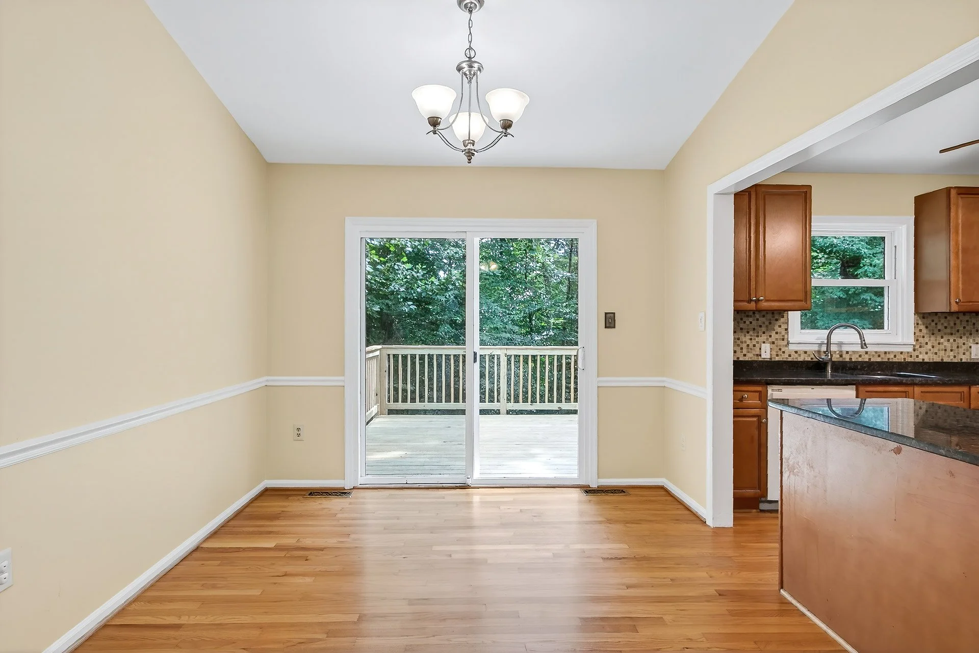 Empty dining area with sliding glass door leading to a wooden deck, visible kitchen area to the right, and hardwood floors.