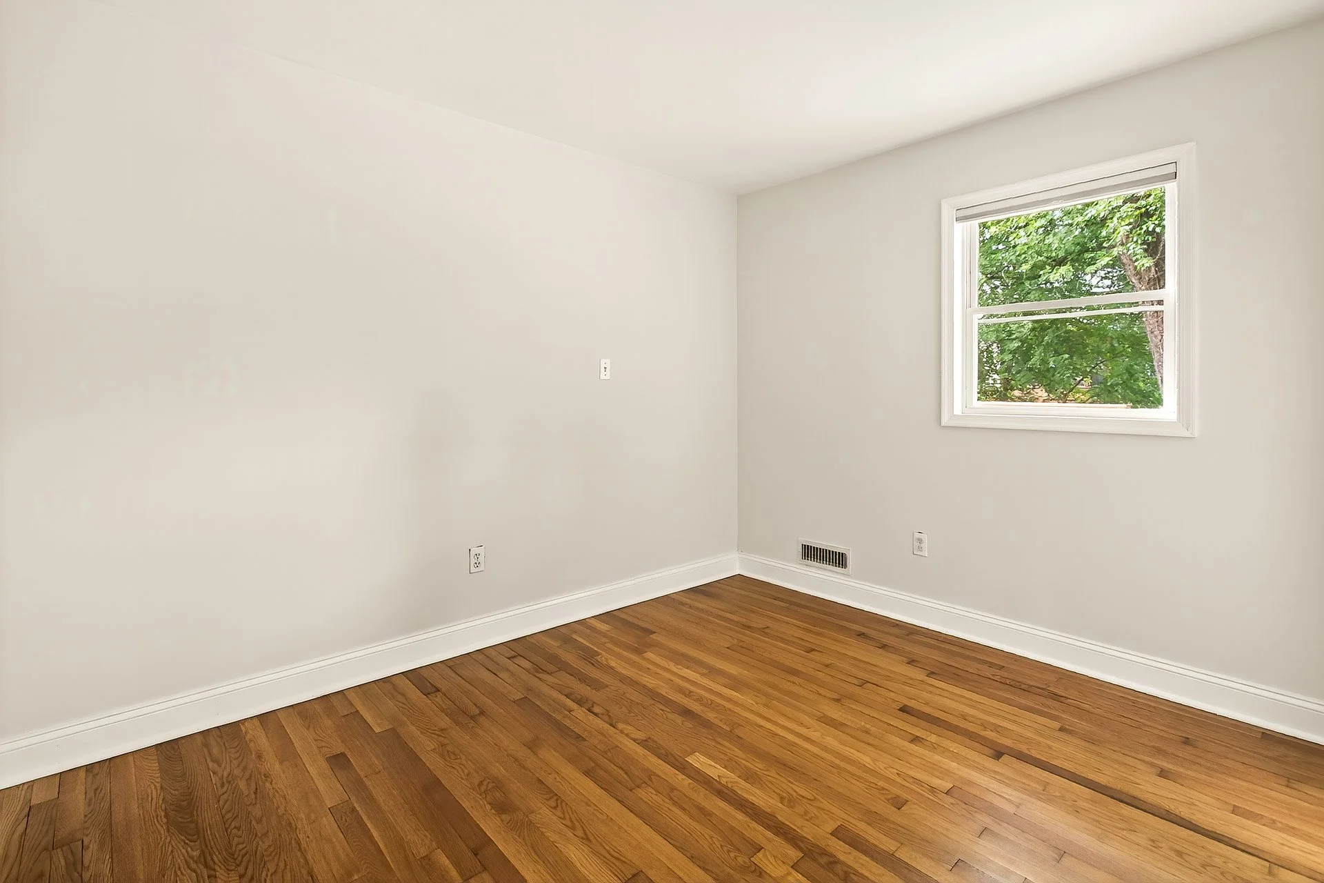 Empty room with a window showing green trees, hardwood floor, white walls, and baseboards.