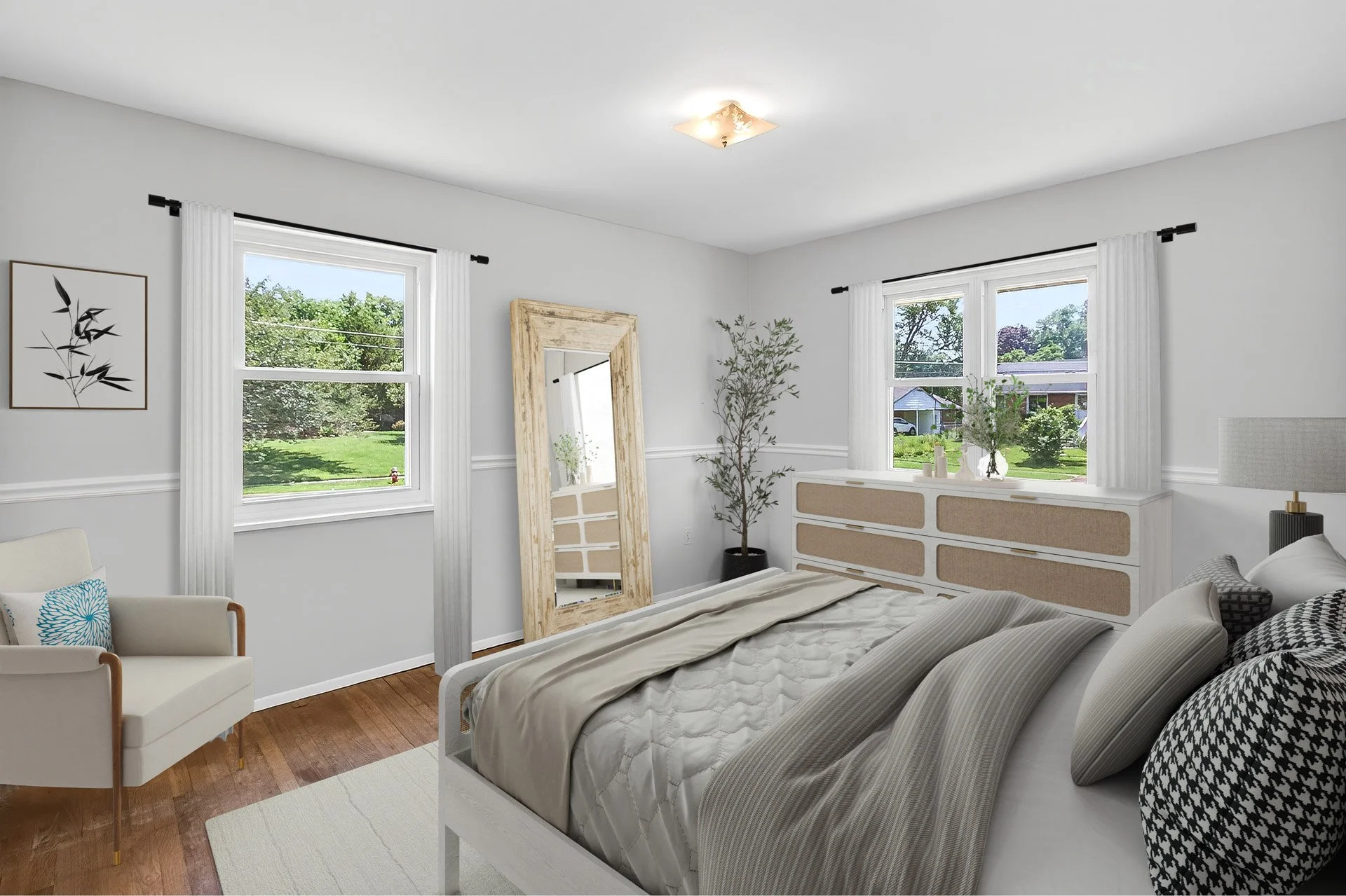 Bright bedroom with white walls and hardwood floor, featuring a bed with beige and black bedding, a large mirror leaning against the wall, a white armchair on the left, and two windows with white curtains showing green trees outside.