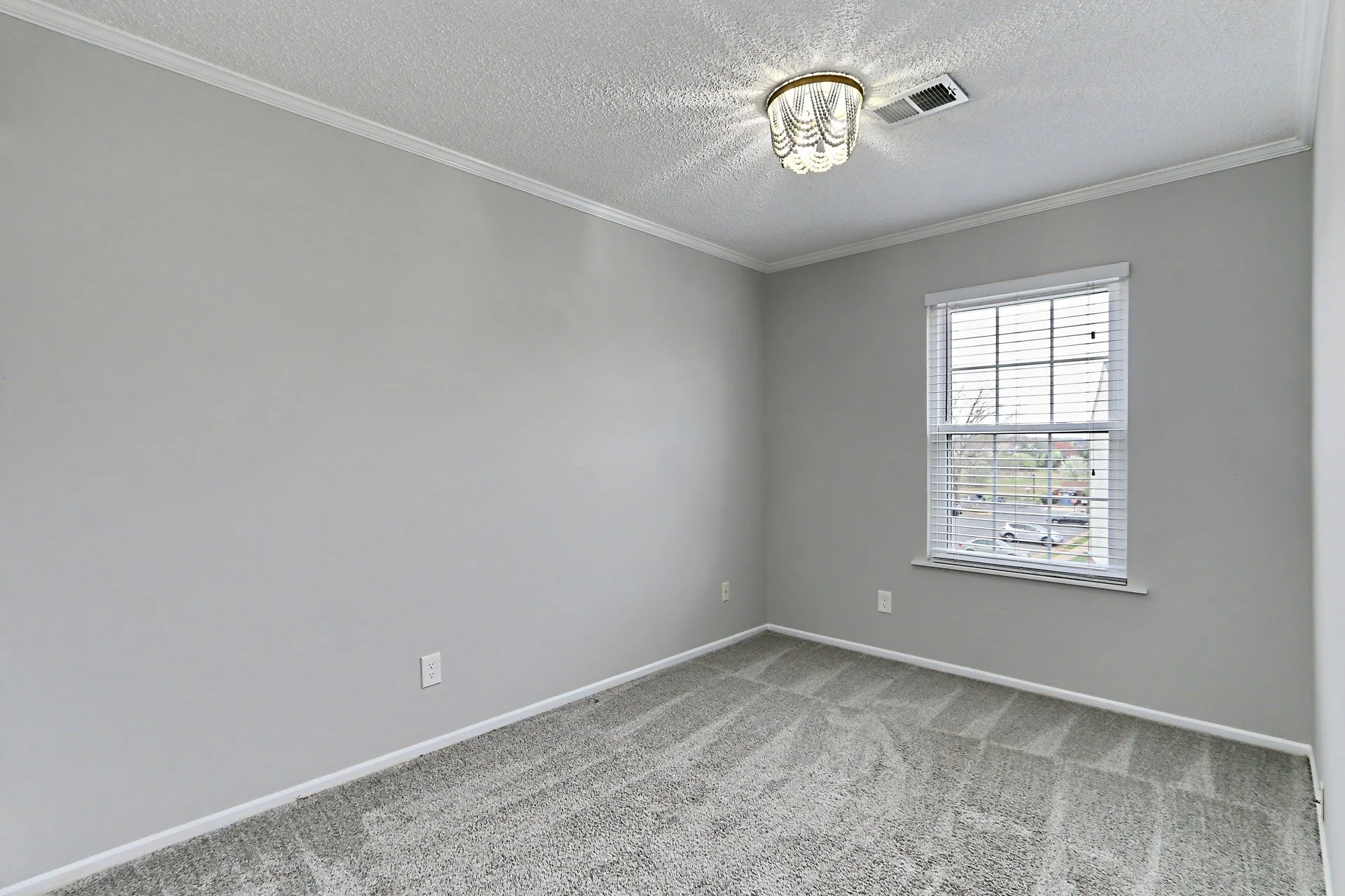An empty room with gray walls, beige carpet, a window with white blinds, a ceiling light fixture, and an air vent.