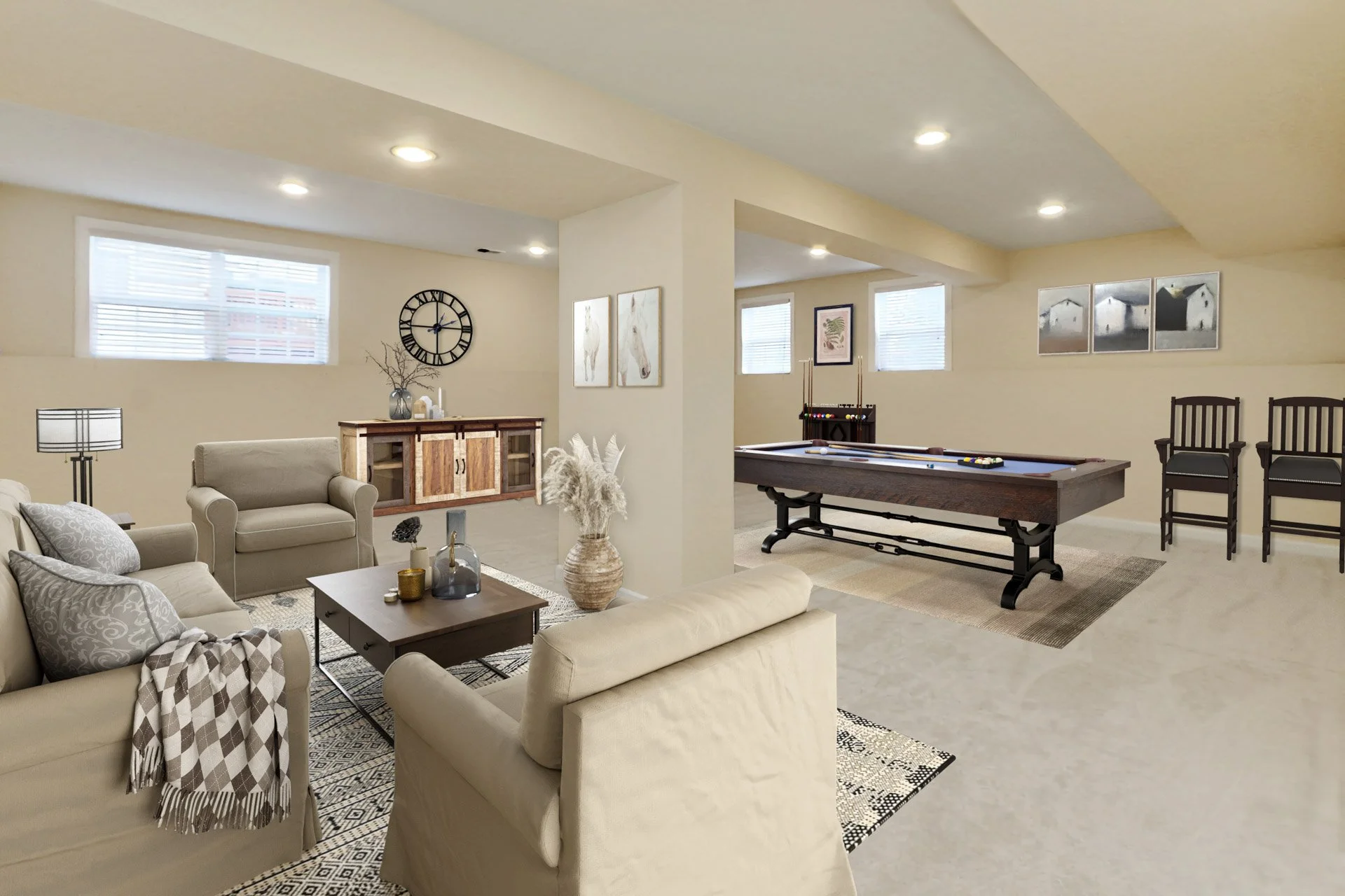 A spacious basement recreation room with a beige sofa, armchair, and a patterned accent chair. There is a dark wood coffee table with decorative vases and candles. A large wall clock hangs above a wooden sideboard. In the background, a pool table is