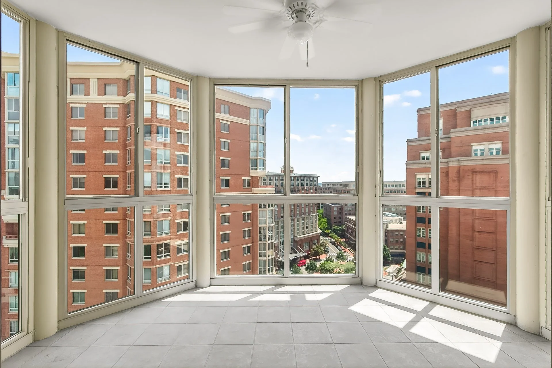 Interior view of a glass-enclosed balcony or sunroom with tiled floor, overlooking a cityscape of tall brick buildings under a blue sky.