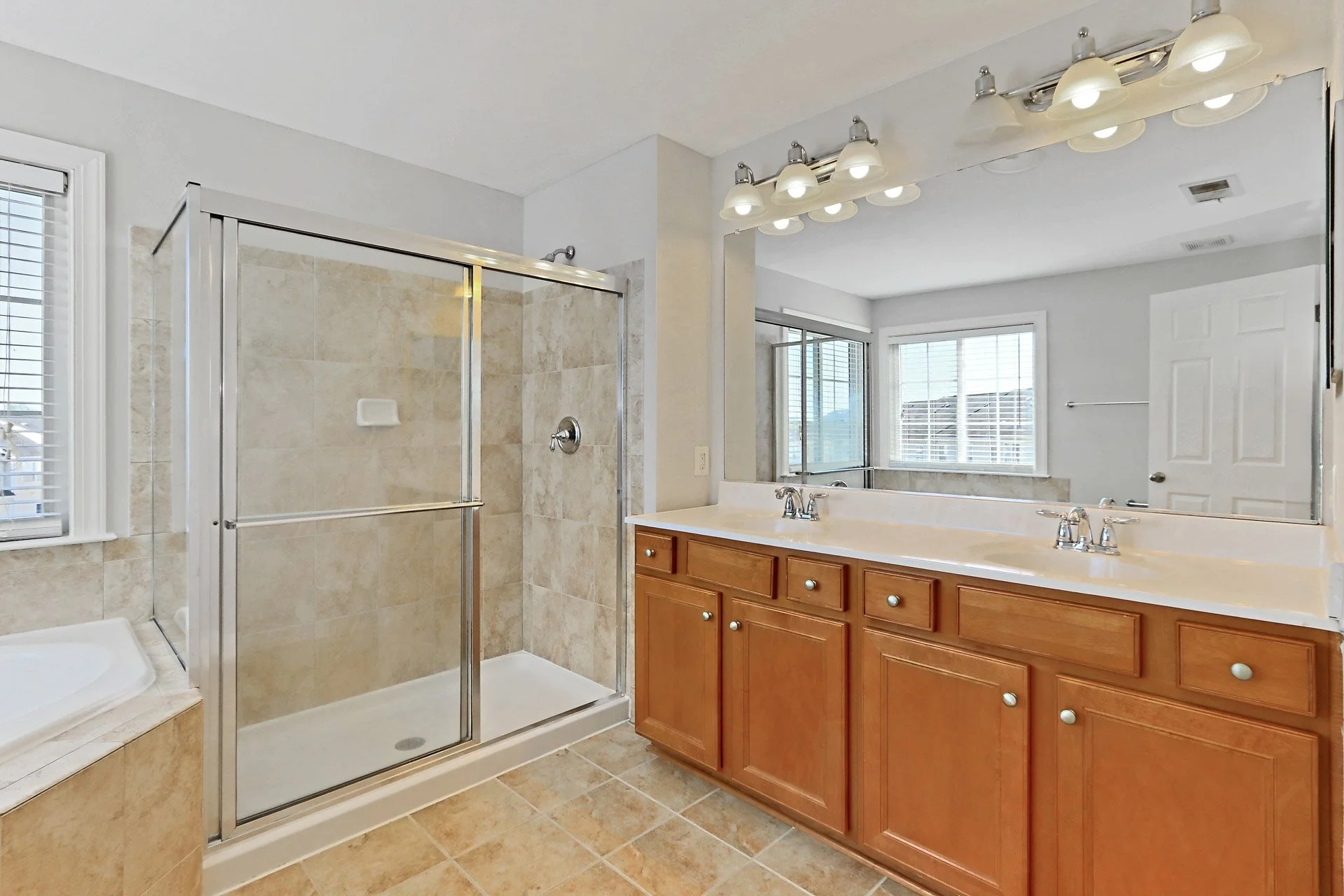 Bathroom featuring a glass-enclosed shower, double sink vanity with wooden cabinets, large mirror, and windows with blinds.