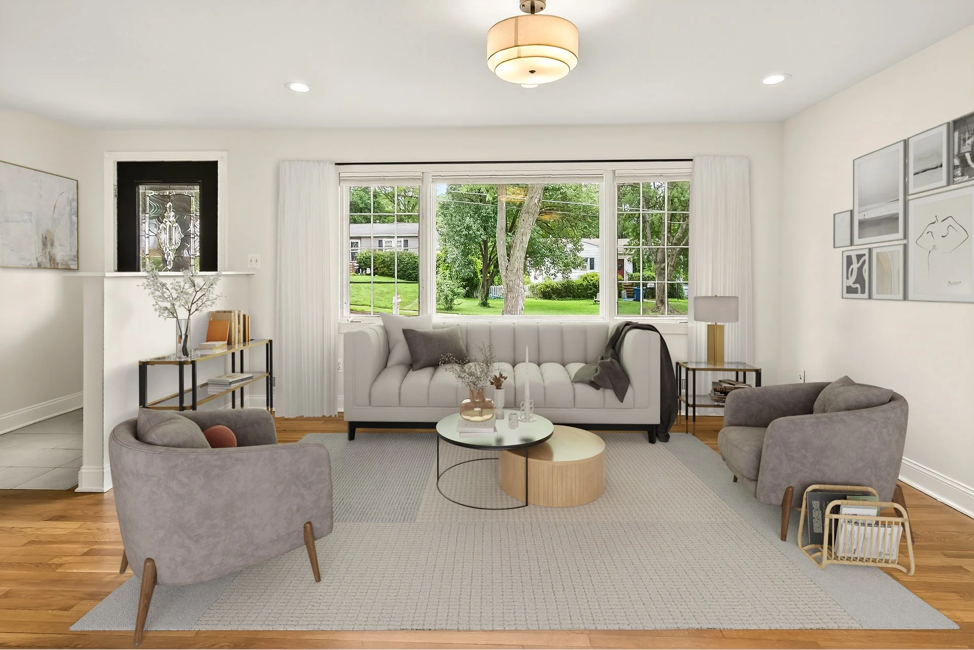 Living room with white walls, large window facing outside, beige rug, gray armchairs, white sofa with gray cushions, round coffee tables, artwork on walls, and a wooden magazine holder.