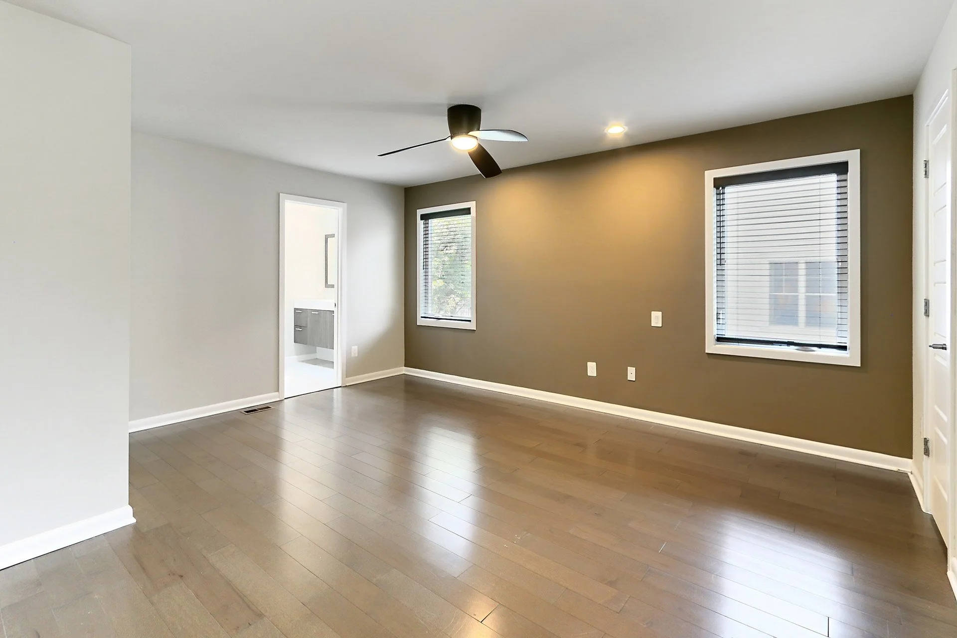 Empty living room with brown and white walls, wood flooring, two windows with blinds, a ceiling fan, and a doorway leading to another room.