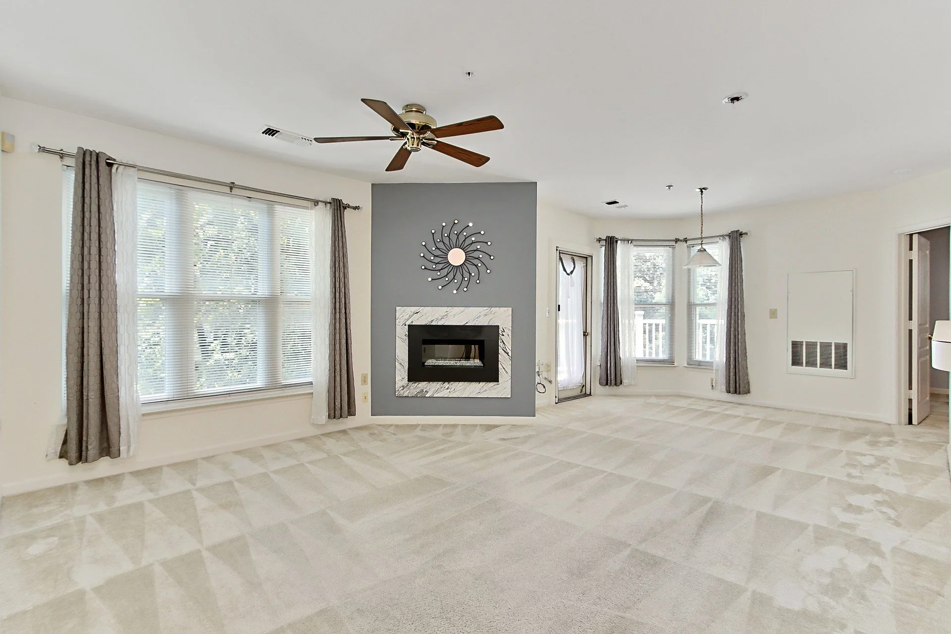 Empty living room with large windows, beige carpet, gray accent wall, modern fireplace, ceiling fan, and natural light.