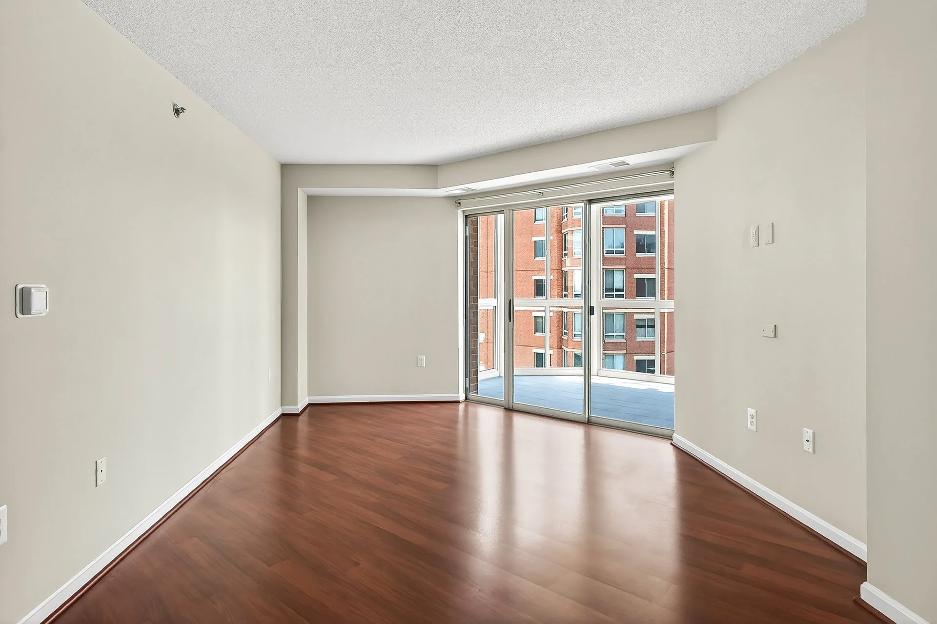 Empty living room with hardwood floors, white painted walls, large glass sliding door leading to a balcony, and an adjacent high-rise brick building outside.