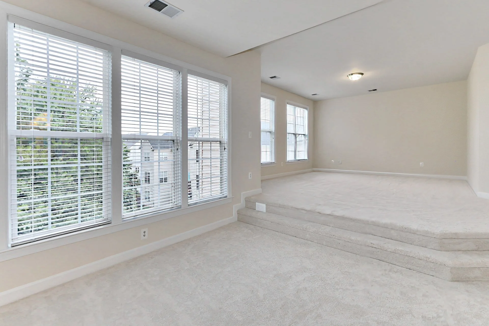 Empty living room with large windows with white blinds and carpeted floor, some steps leading up to a slightly elevated area, beige walls, and ceiling lights.