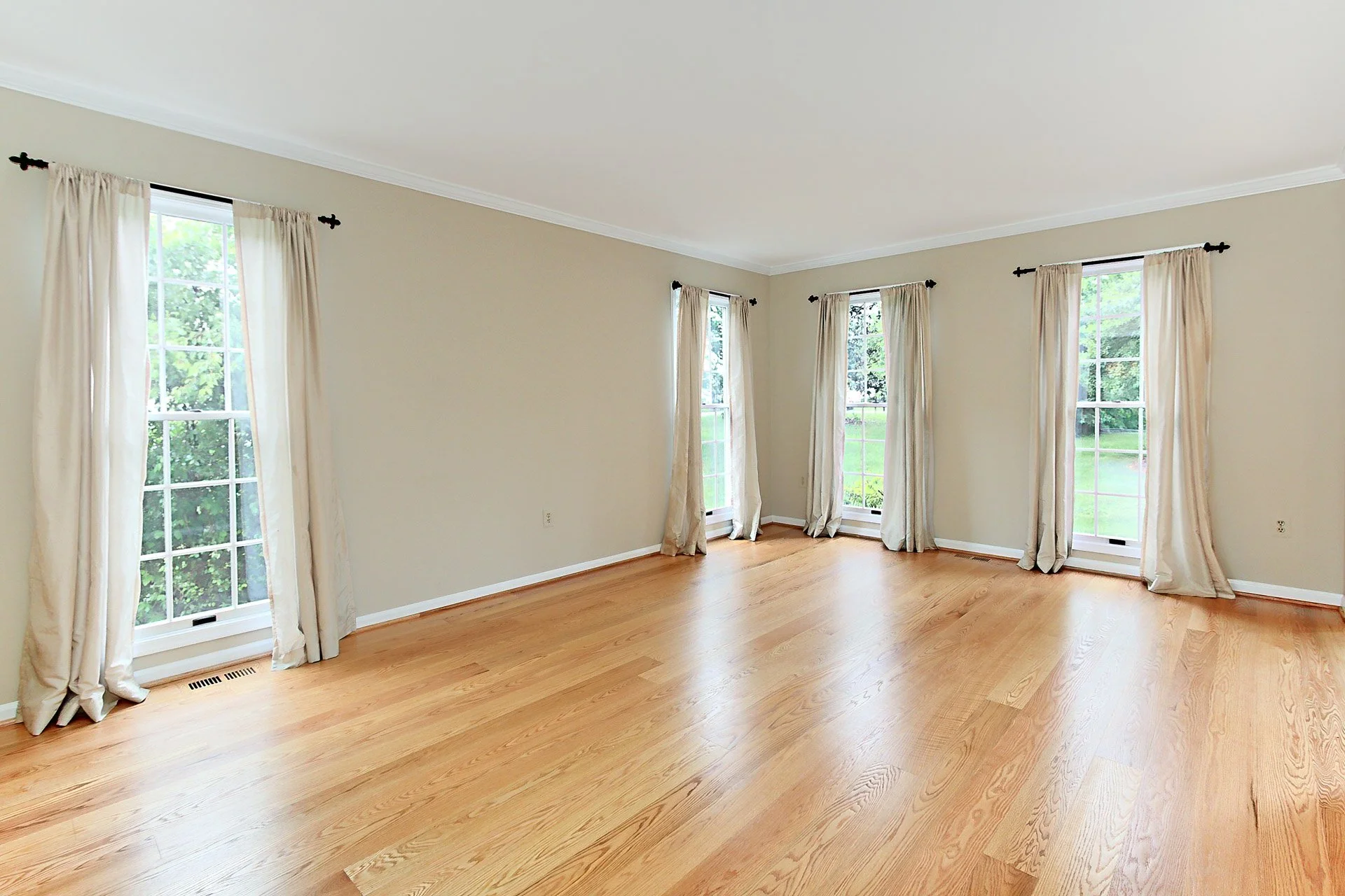 Empty living room with hardwood floors, four large windows with beige curtains, and cream-colored walls.