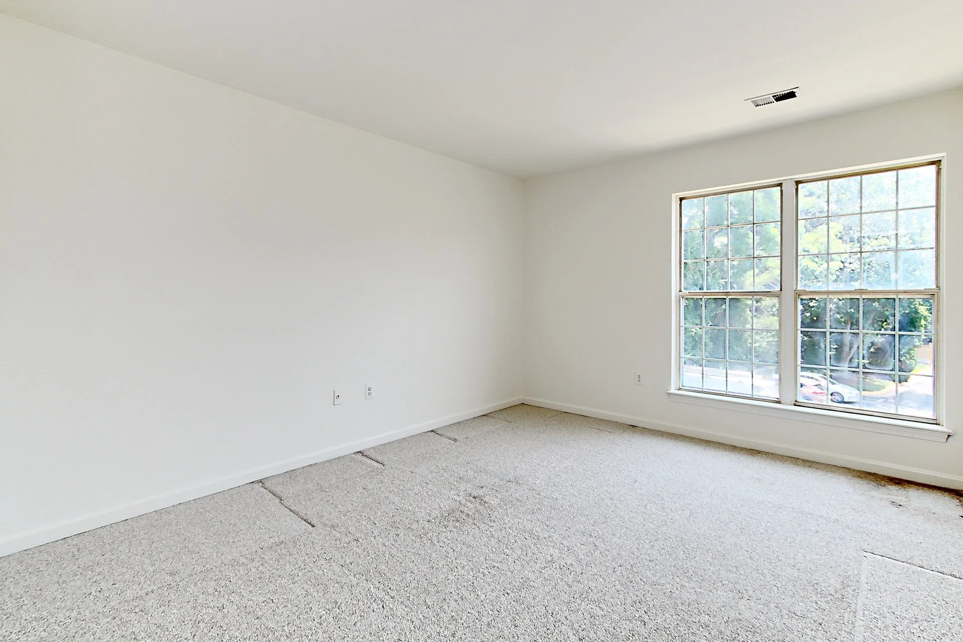 Empty room with white walls, beige carpet, large double window showing trees outside, ceiling vent, and electrical outlets.