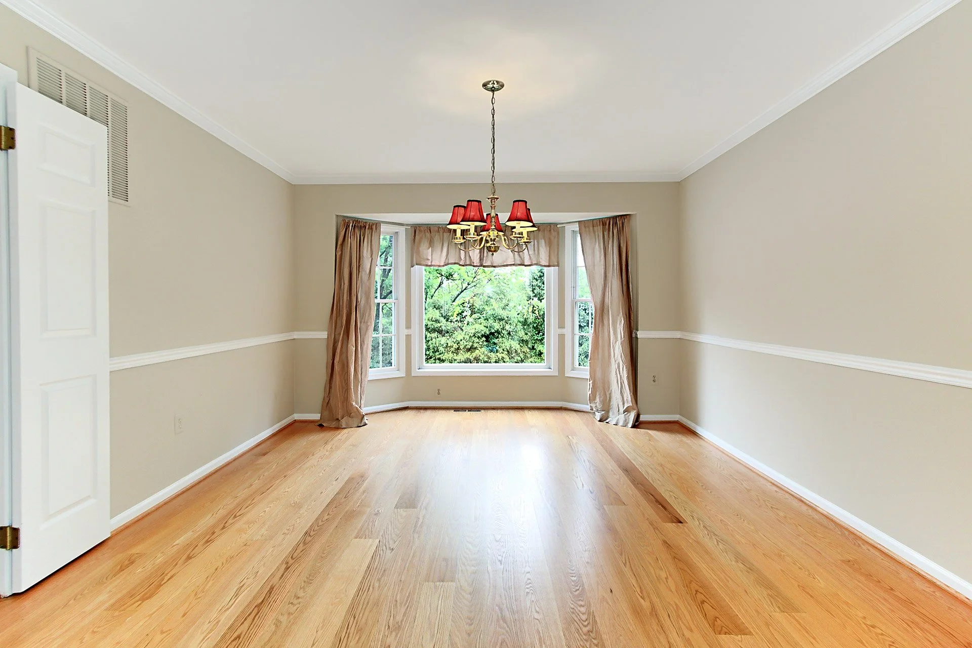 Empty room with hardwood floors, beige walls, white trim, a bay window with curtains, and a chandelier.