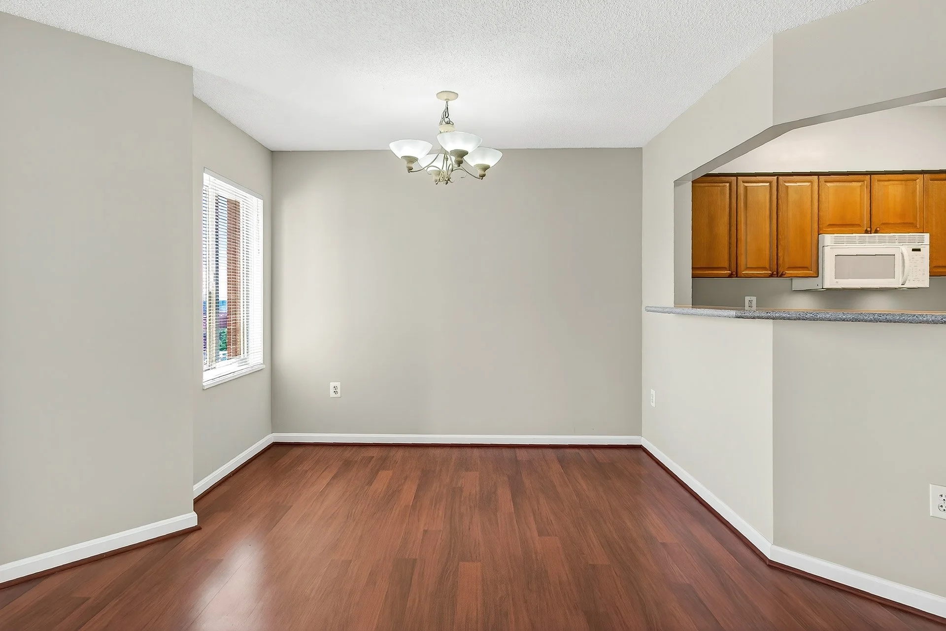 Empty dining area with hardwood floors, a window with blinds, a ceiling chandelier, and a view into the kitchen with wooden cabinets and white appliances.