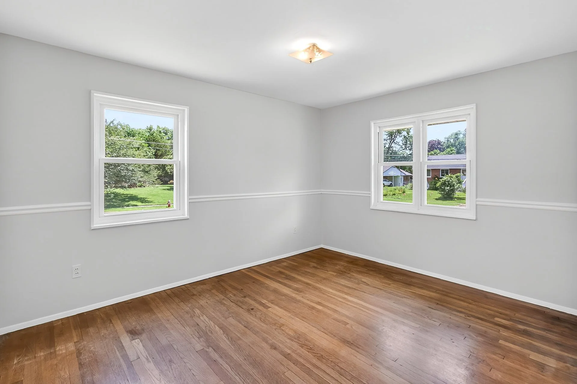 Empty room with white walls, hardwood floors, and three double-hung windows showing a green lawn outside.
