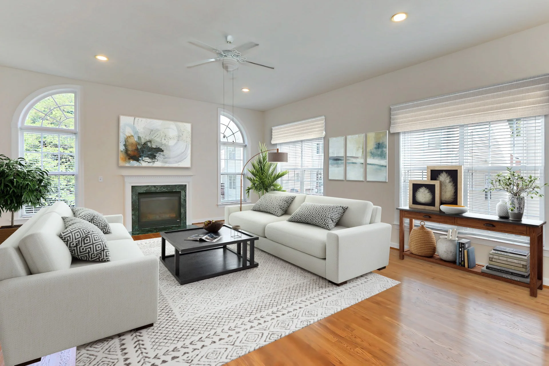Modern living room with white sofas, a black coffee table, a fireplace, large windows with blinds, artwork on the walls, a potted plant, and decorative items on a wooden console table.