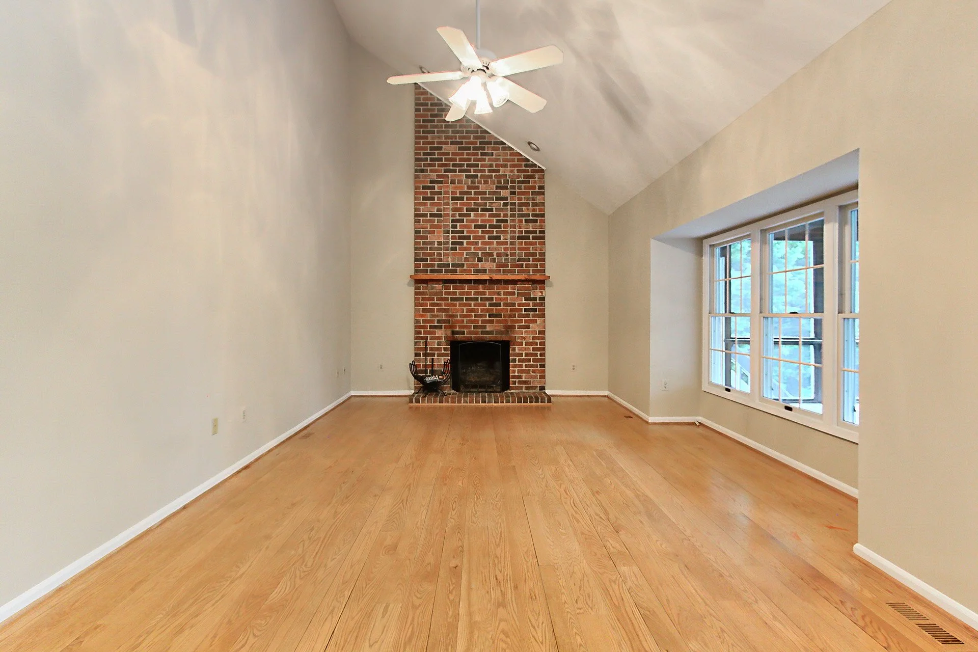 Empty living room with hardwood floors, a brick fireplace, large windows, and a ceiling fan.