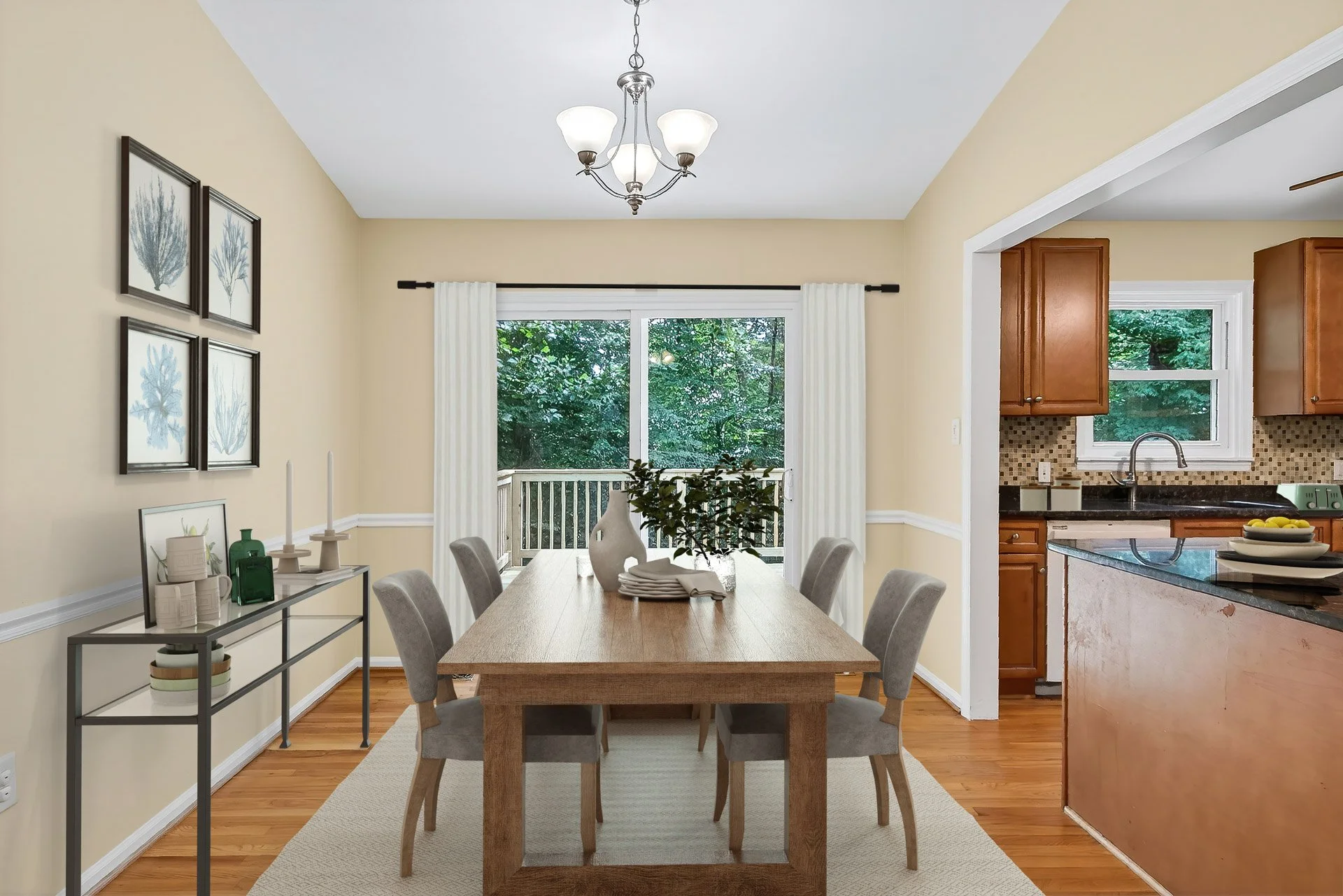 Dining room with beige walls, a wooden table with six gray upholstered chairs, a sliding glass door with white curtains leading to a porch with trees outside, a black metal console table with decorative items, a chandelier with five lights, and a kit