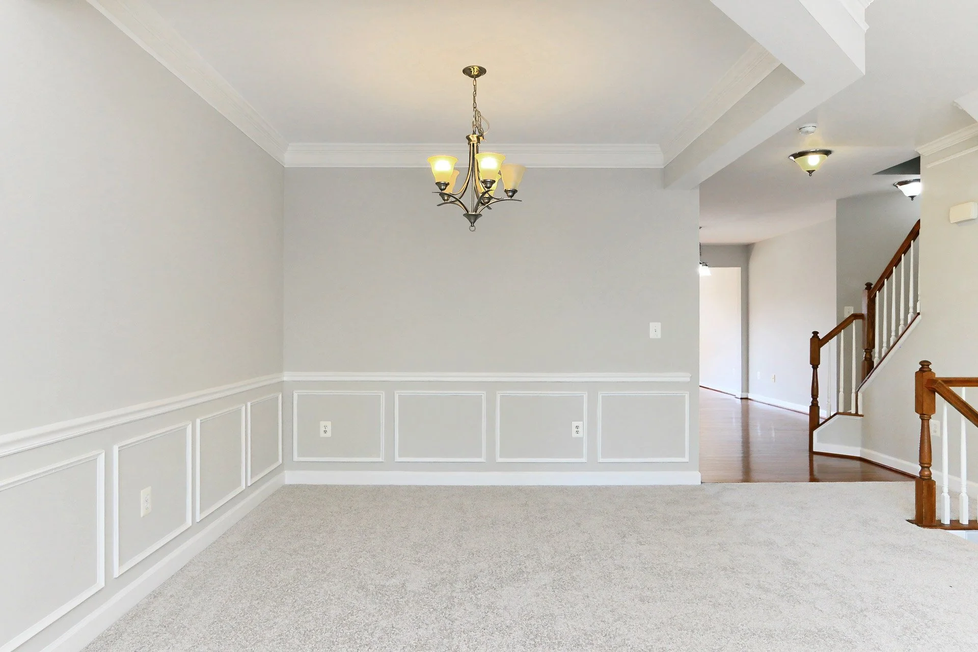 Empty living room and dining area with beige carpet, white walls with wainscoting, a chandelier, and a staircase with wooden handrails.