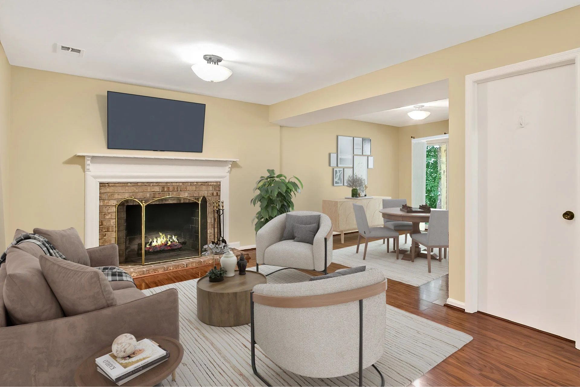 Living room with beige walls, a brick fireplace with a white mantel, a flat-screen TV above, and furniture including sofas, chairs, a round coffee table, and a dining area in the background. There's a large potted plant near the fireplace and a windo