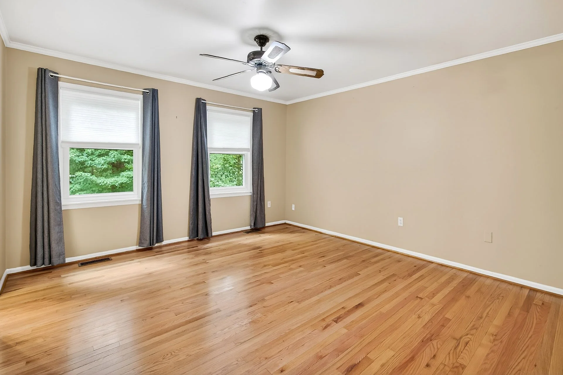 Empty room with hardwood floors, beige walls, two windows with white blinds and dark curtains, ceiling fan with light.