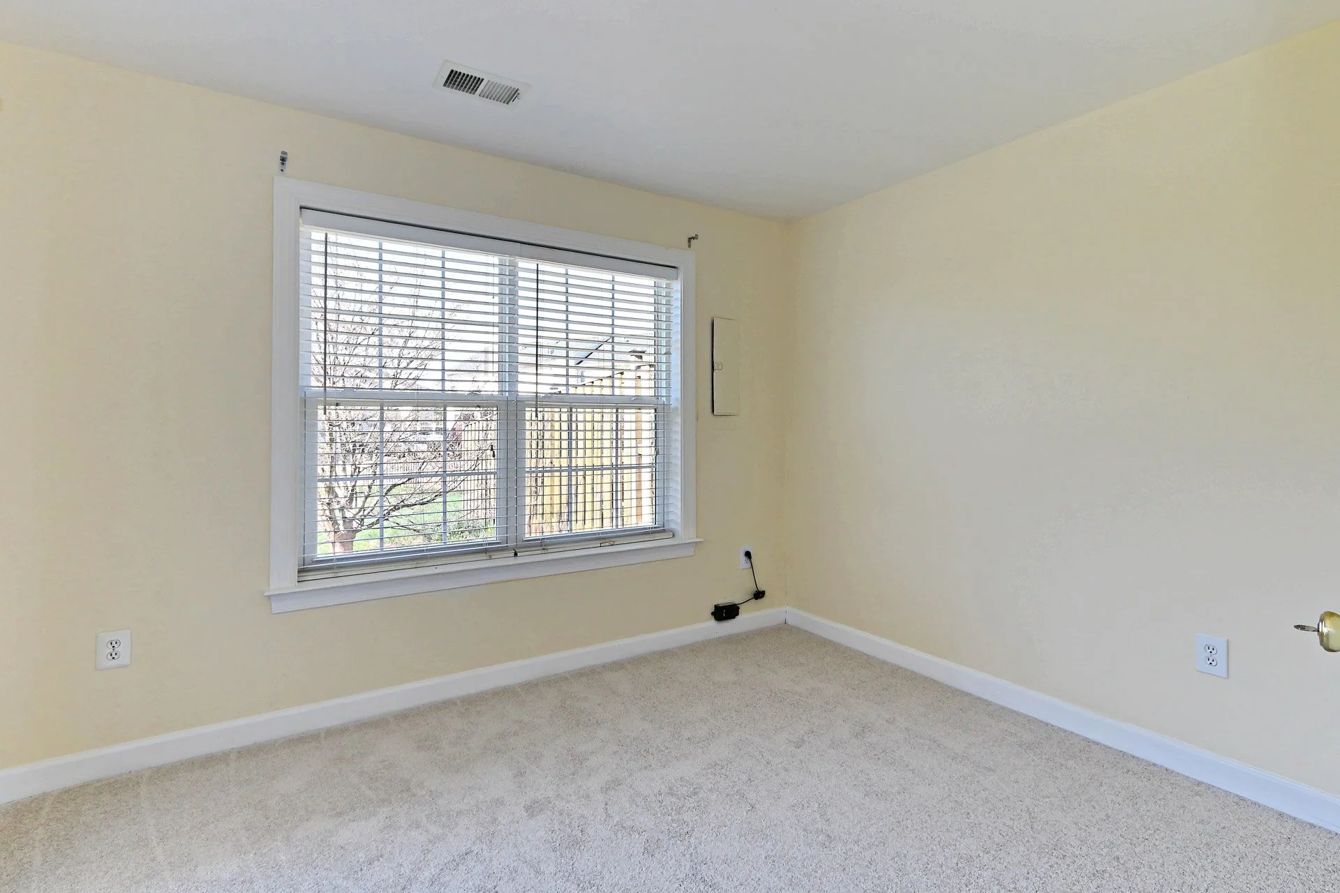 Empty room with a large window, beige walls, carpeted floor, and some electrical outlets and wires.