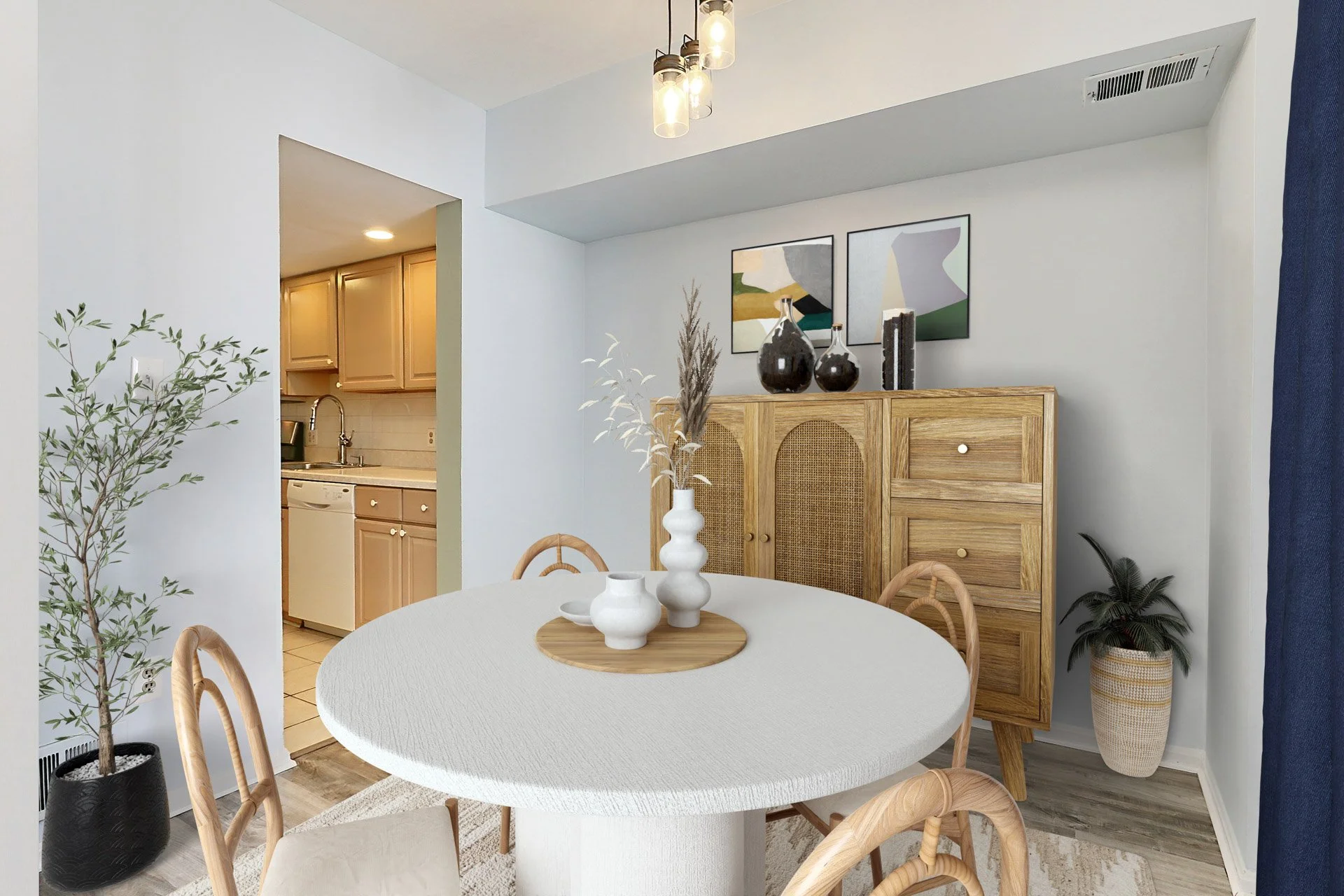 Dining room with white round table, light wooden chairs, and decorative vases on the table. A wooden sideboard with art and vases on top is against the wall. Nearby, there is a potted plant and a painted wall art piece. The background shows a kitchen