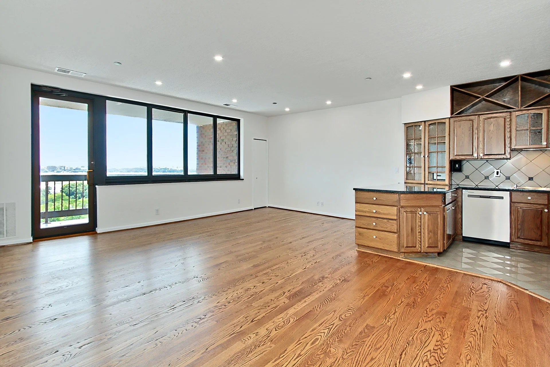 Empty living room with large window, wood floors, and kitchen with wooden cabinets.