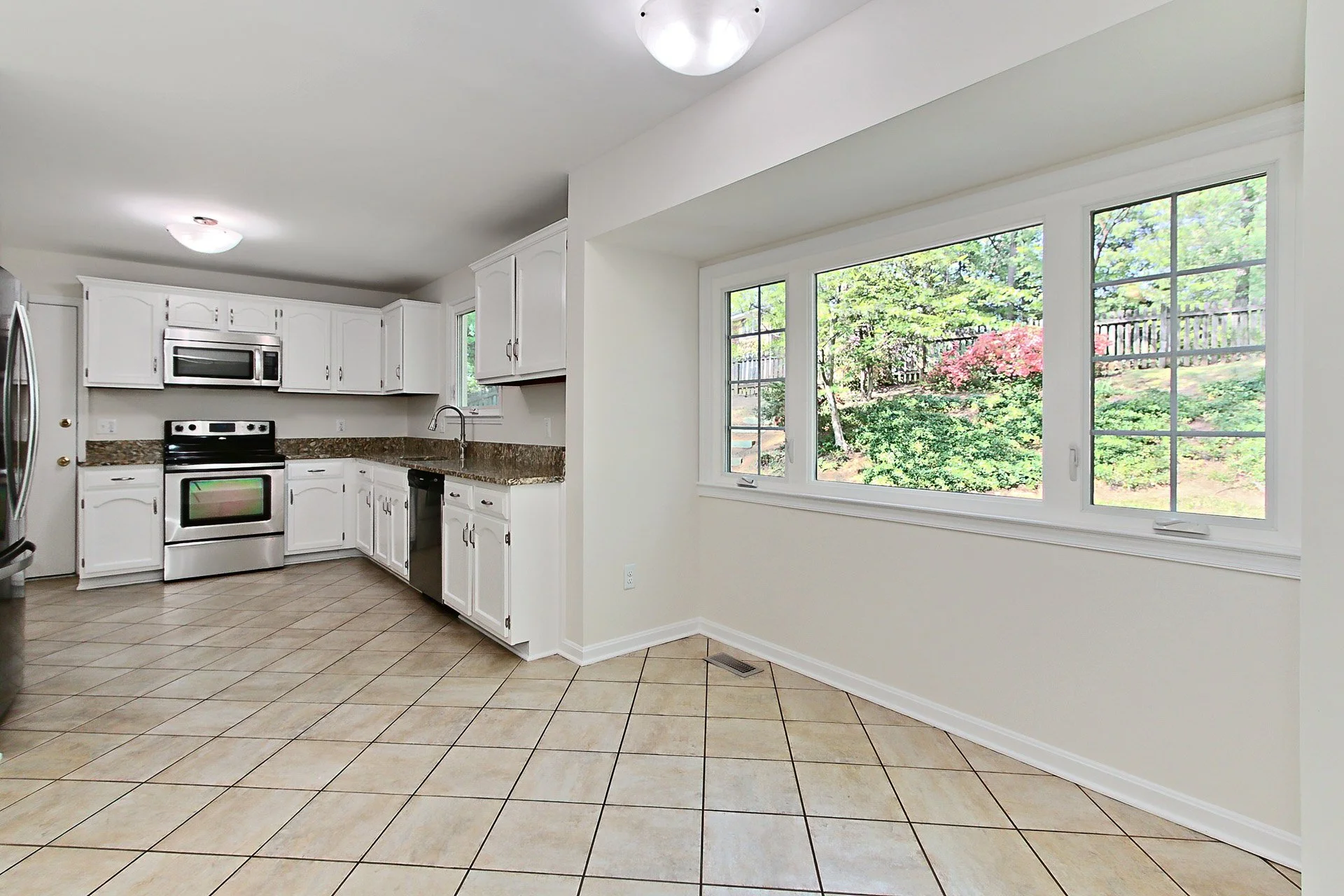 Kitchen with white cabinets, granite countertops, stainless steel appliances, tiled floor, and a window showing a backyard with trees and shrubs.
