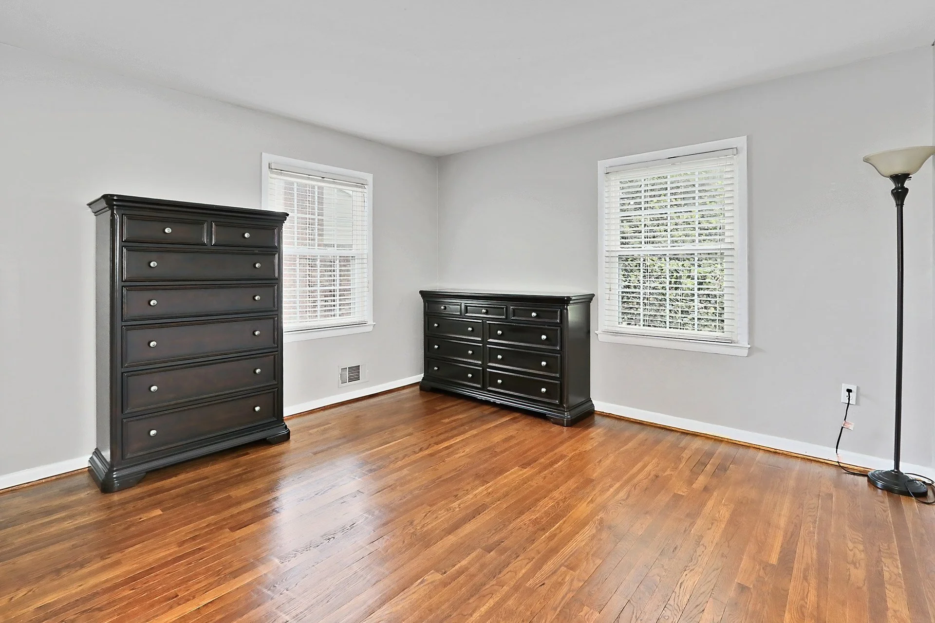Empty bedroom with hardwood floors, two windows with white blinds, two black dressers, and a tall floor lamp.