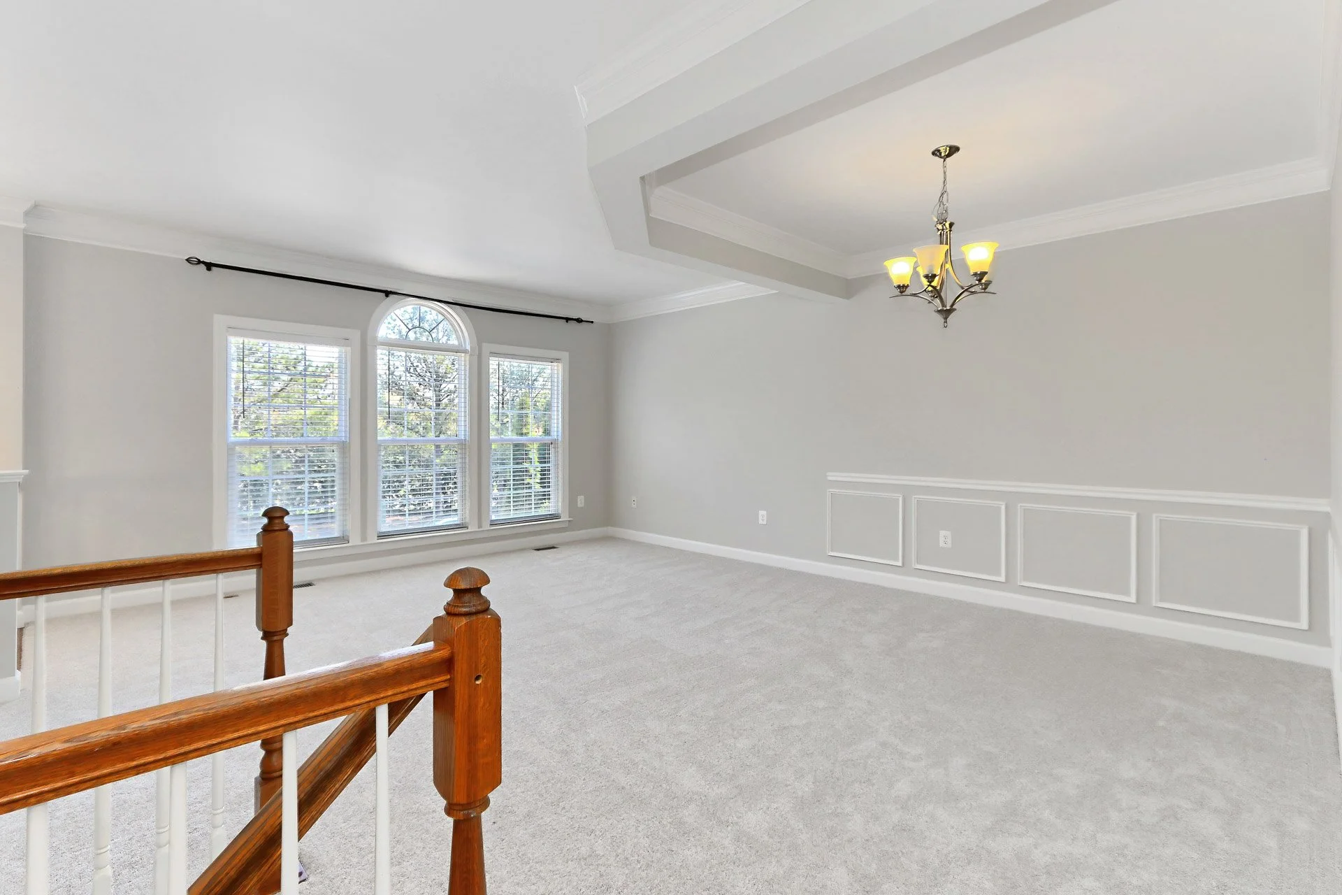 Empty living room with multiple windows, chandelier, light gray walls, white crown molding, and beige carpet.