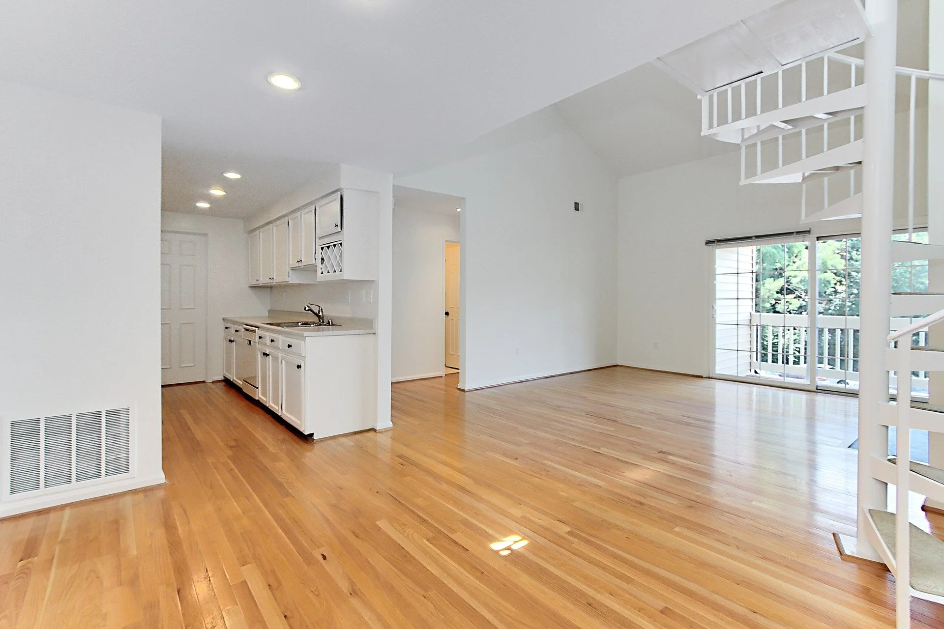 Empty living room with hardwood floors, white walls, large window with blinds, and a spiral staircase leading to an upper level. Kitchen area with white cabinets and a small sink.