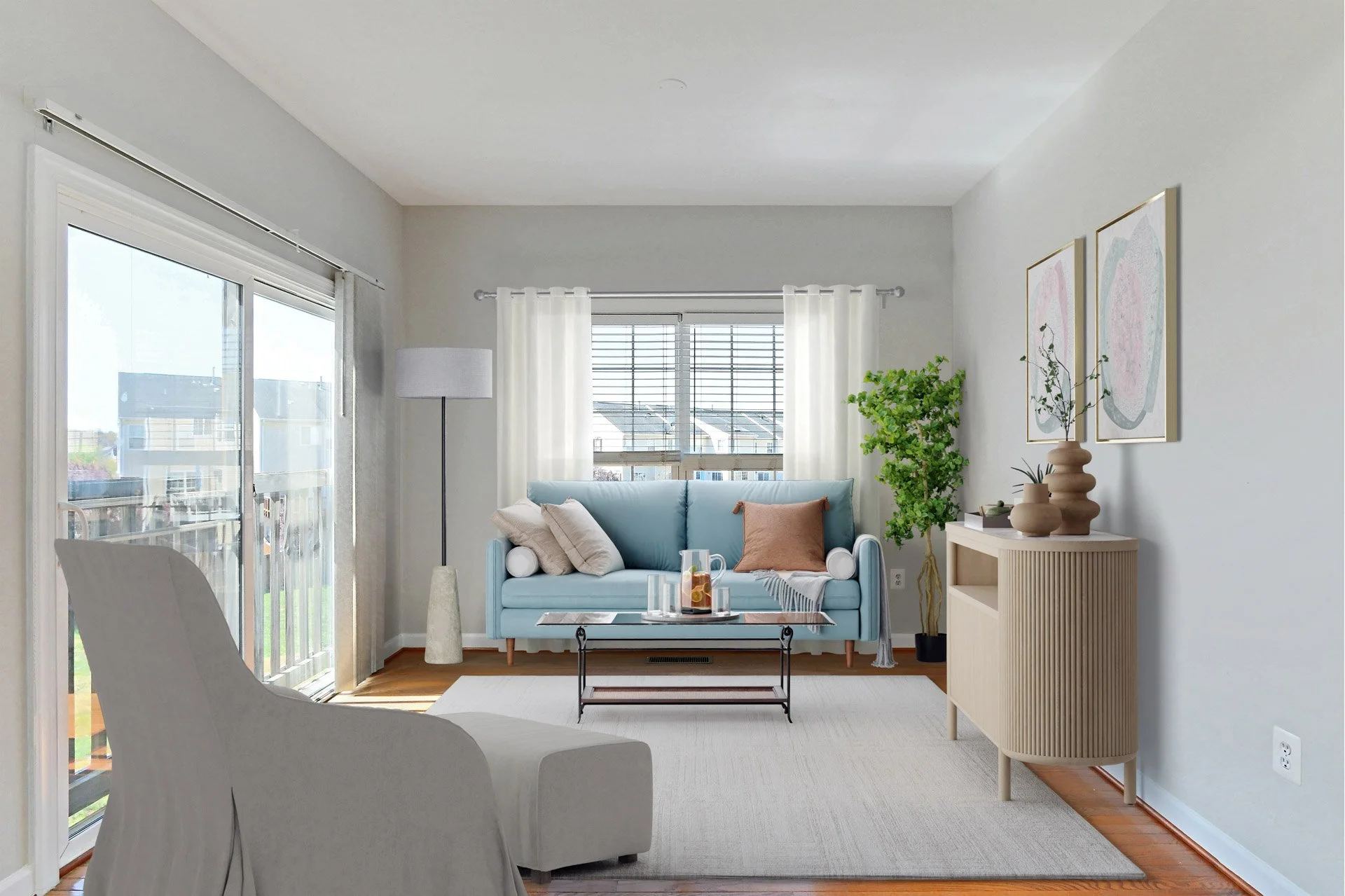 Bright living room with large window, light blue sofa, white curtains, decorative pillows, modern coffee table, potted plant, framed artwork, light-colored sideboard, and white armchair.